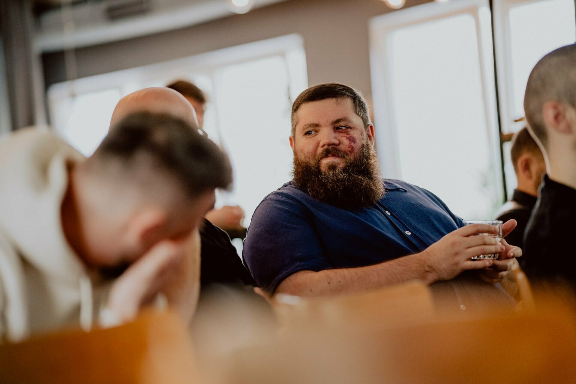 A bearded man in a blue shirt sits at a table with a drink in his hand and smiles at another man in a white hooded sweatshirt who covers his face with his hand. Several other people blur in the background, creating a warm and social atmosphere reminiscent of an informal press meeting. 