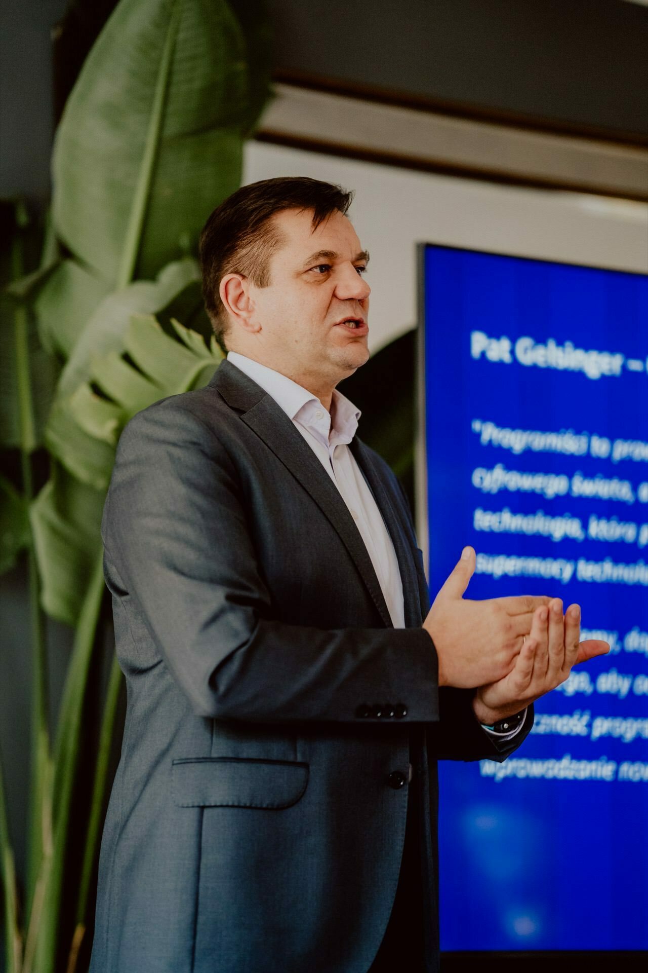 A man in a dark suit and white shirt is speaking in front of a large screen, with a text reminiscent of a press meeting. He holds his hands together as if to emphasize a point, with large green leaves of a plant in the background, creating a professional or corporate atmosphere. 