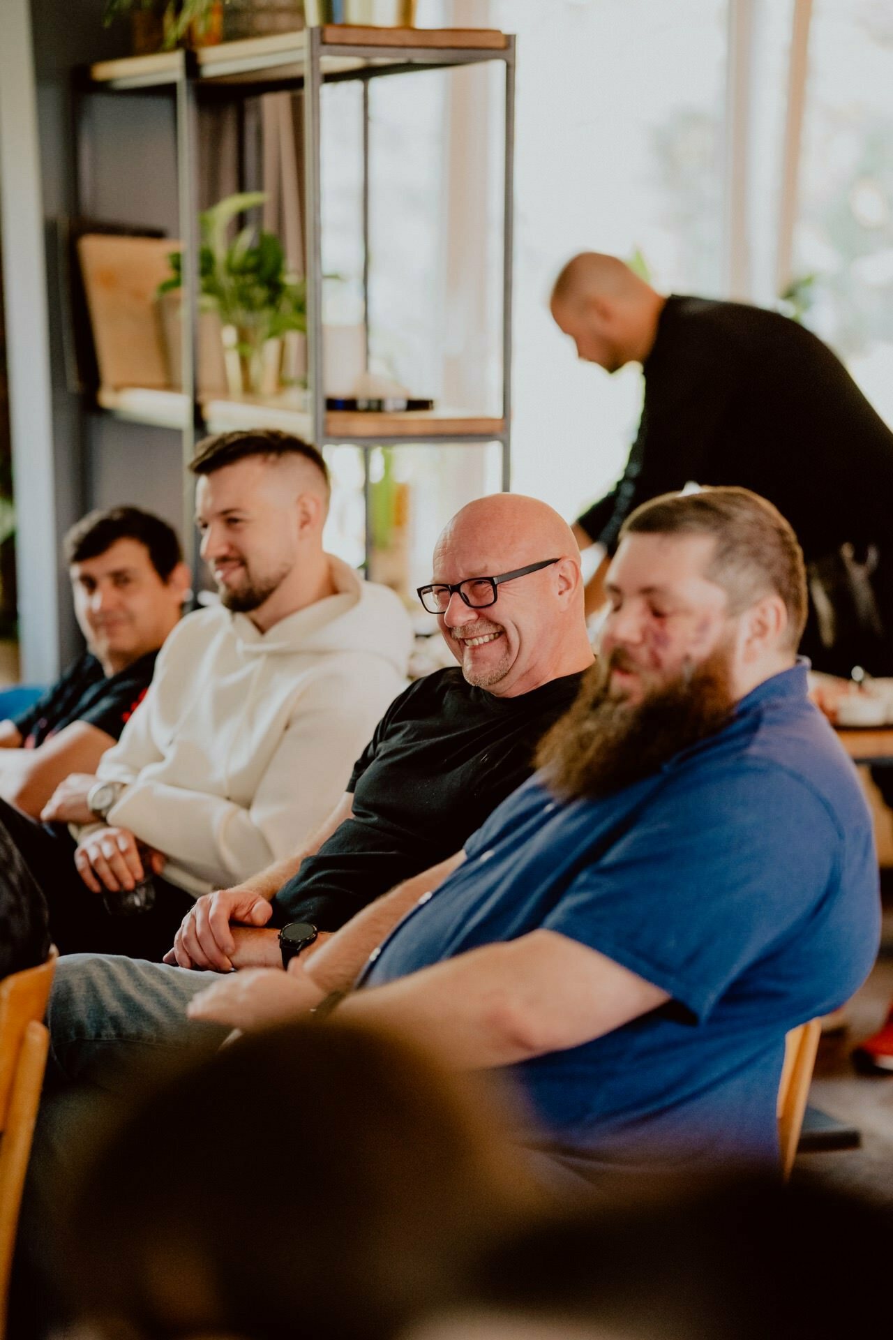 A group of four men sitting in a room, smiling and engaged in conversation. One man wearing glasses and with a bald head appears to be laughing. Another man in the background is bent over and probably working or interacting with someone outside the frame during what appears to be an informal press meeting. They are in a cozy room full of plants.   