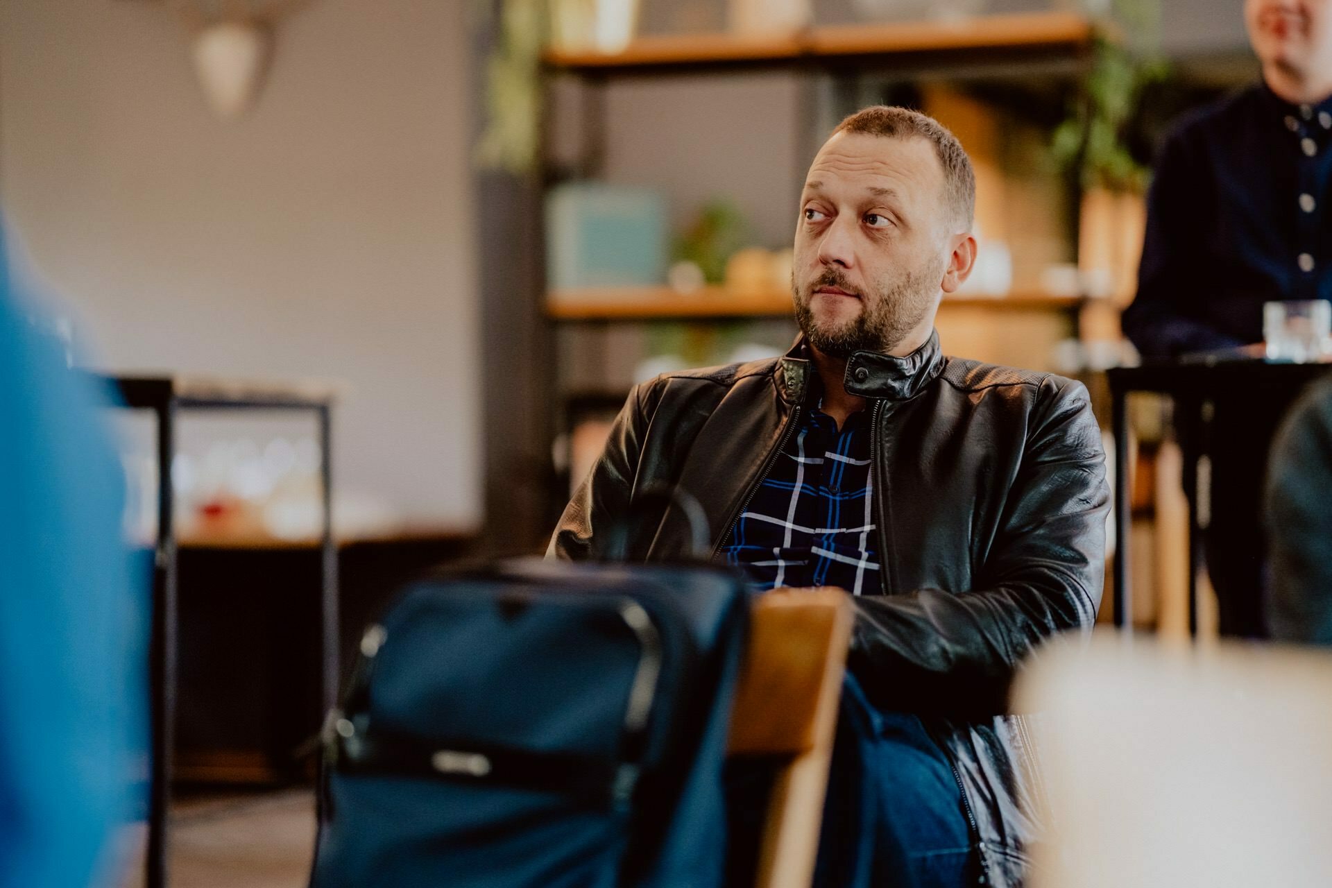 A person with short hair, wearing a leather jacket and a blue plaid shirt, sits attentively in a setting reminiscent of a press meeting, with shelves and plants in the background. On a chair or table next to them lies a blue backpack. 