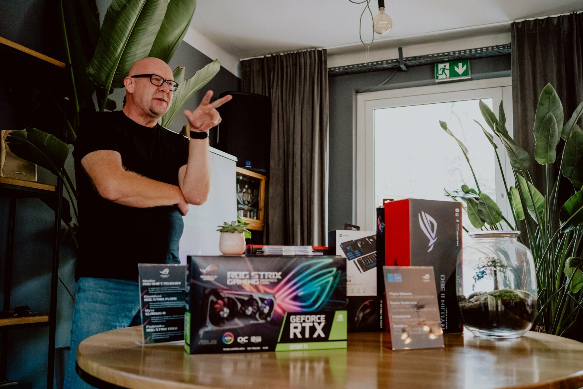 A man stands behind a table with a variety of computer equipment, including a GeForce RTX graphics card, in a room with large leafy plants. As he speaks, he gestures with one hand to suggest that he is explaining or presenting issues at what looks like a press meeting. 