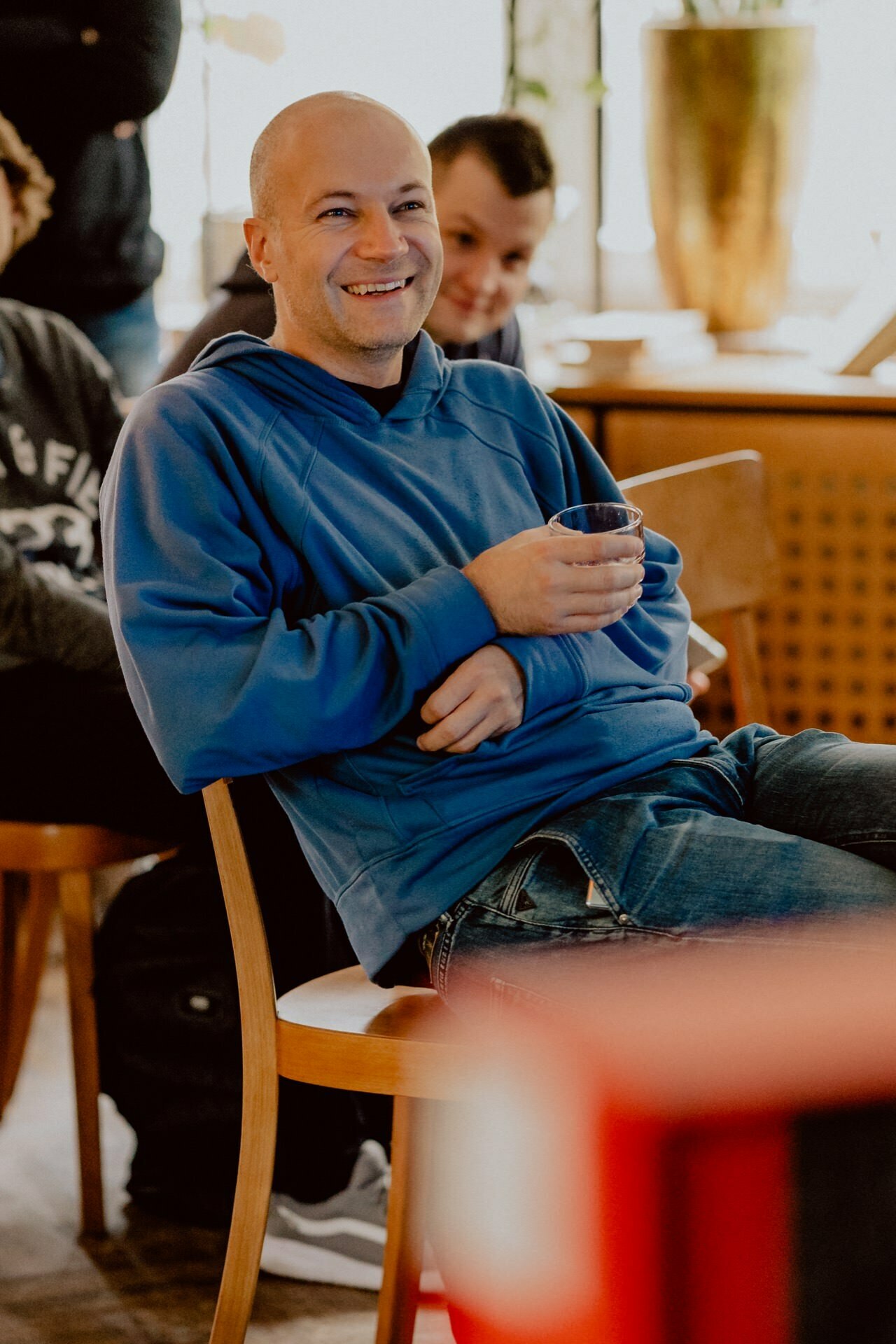 A smiling man with a shaved head, wearing a blue hooded sweatshirt, sits casually on a wooden chair and holds a drink. Another person, also smiling, is partially visible in the background. The setting looks relaxed and informal, almost like an impromptu press meeting.  