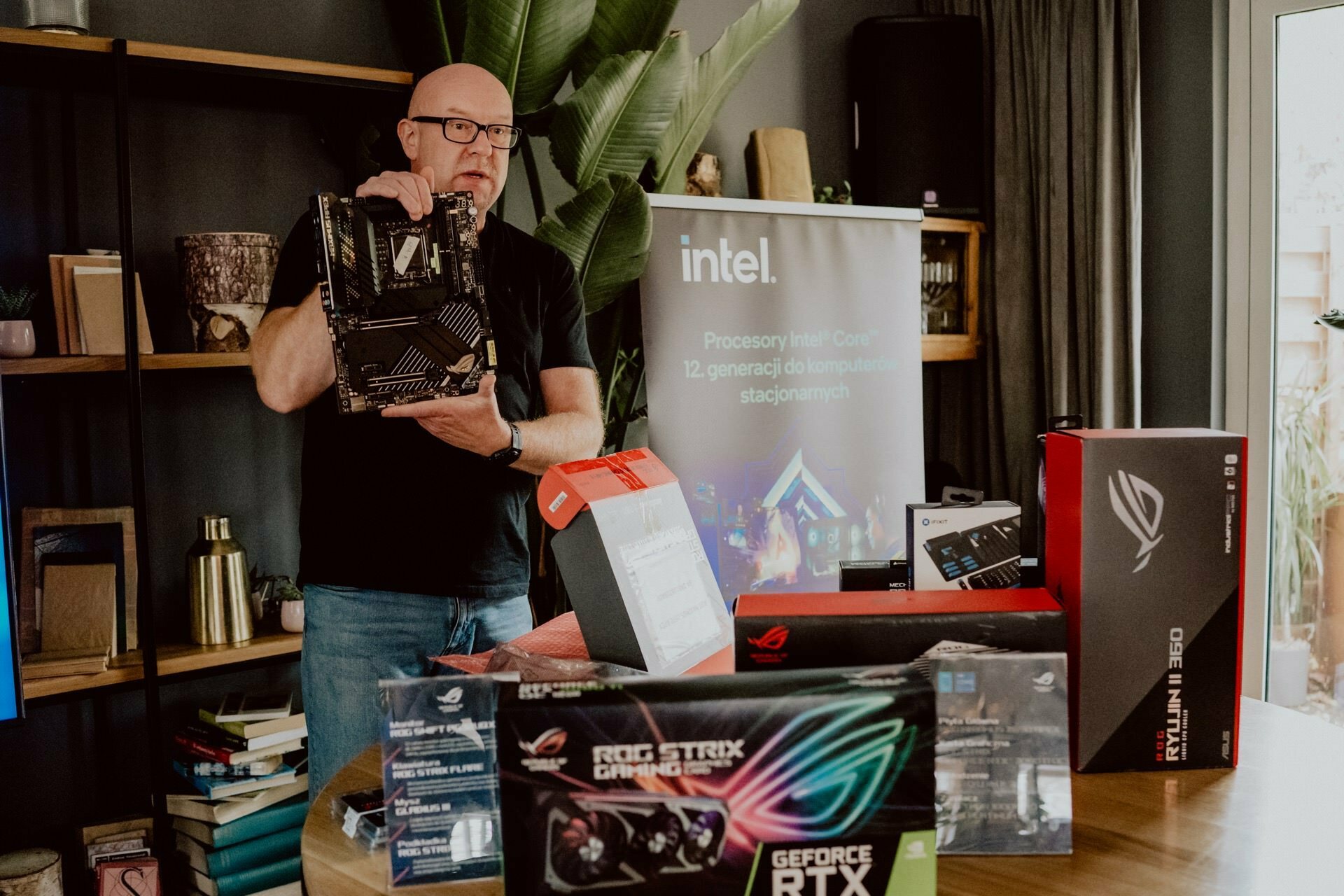 A man stands in a room, holding a computer motherboard, and various computer components and packaging on a table in front of him. A large "interview" banner is partially visible in the background, suggesting that this is a press meeting. The room is decorated with plants and bookshelves.  