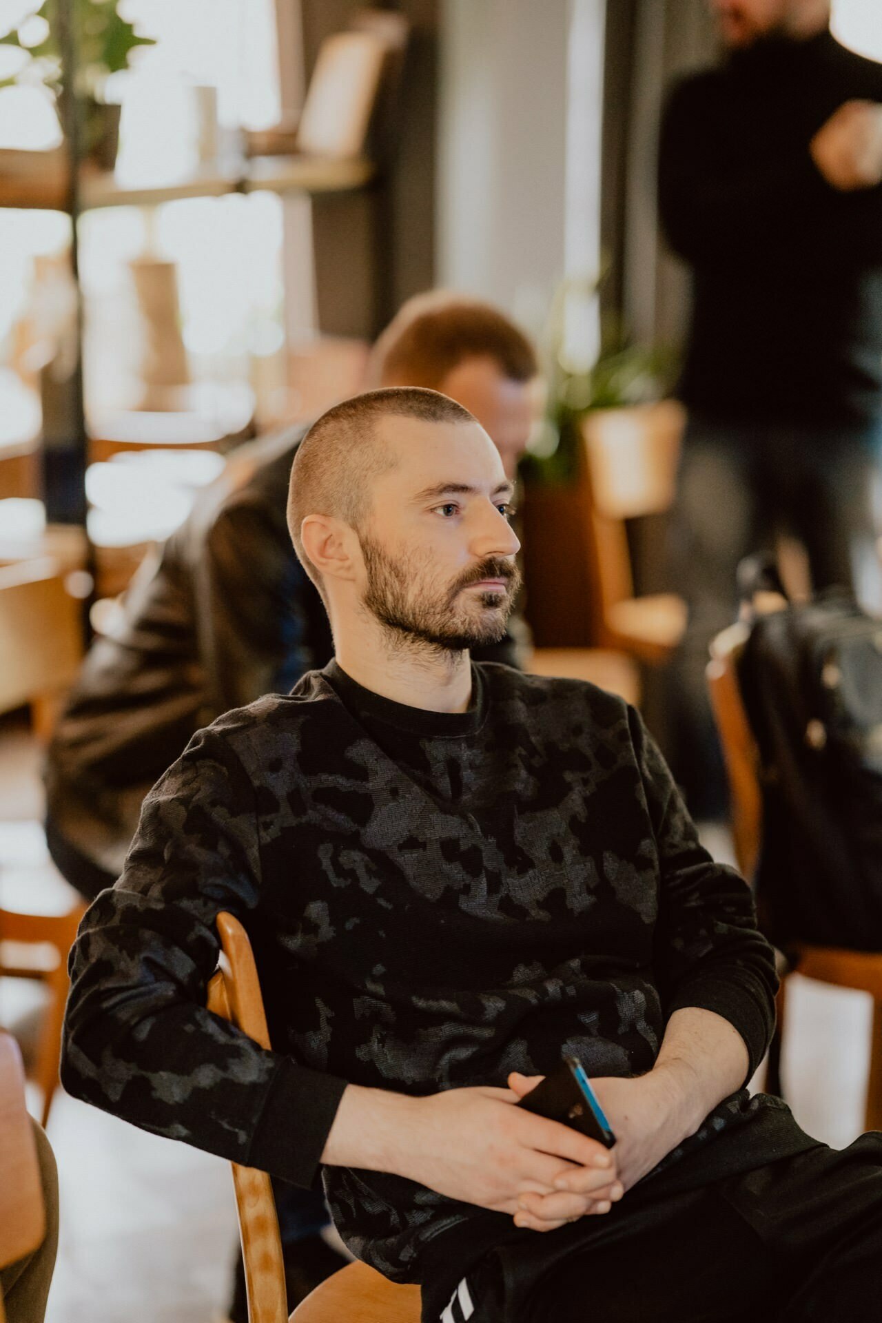 A man with a shaved head and beard sits on a wooden chair in a cozy, well-lit room, perhaps preparing for a press meeting. He is wearing a dark, patterned long-sleeved shirt and holding a phone in his hands. Two other people are visible in the background, somewhat out of focus.  