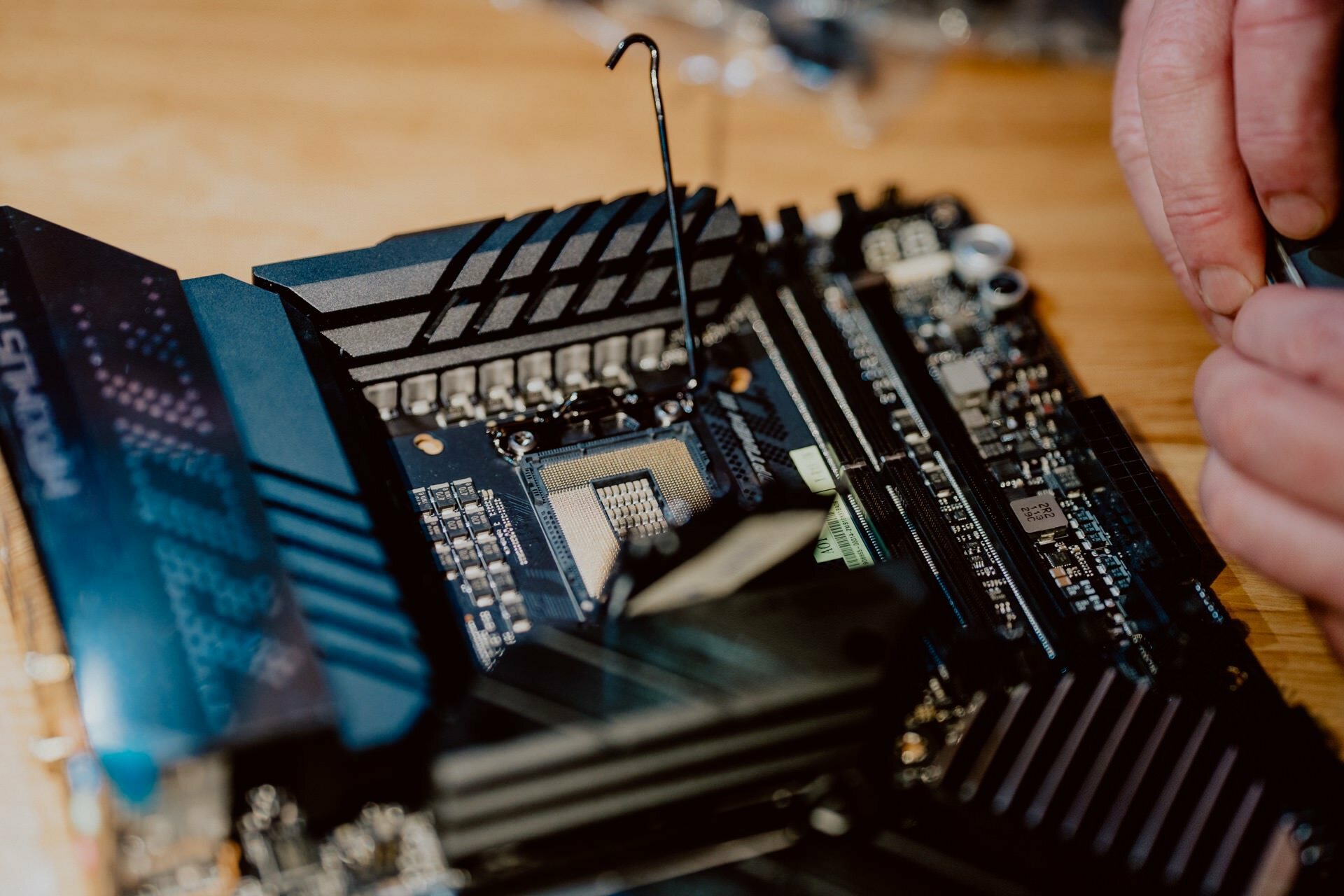 Close-up of hands working on a computer motherboard, with special attention to the processor socket. A metal lever is lifted, preparing to install or remove the processor. The surface, reminiscent of hardware seen at a press briefing, is littered with various components and connectors.  