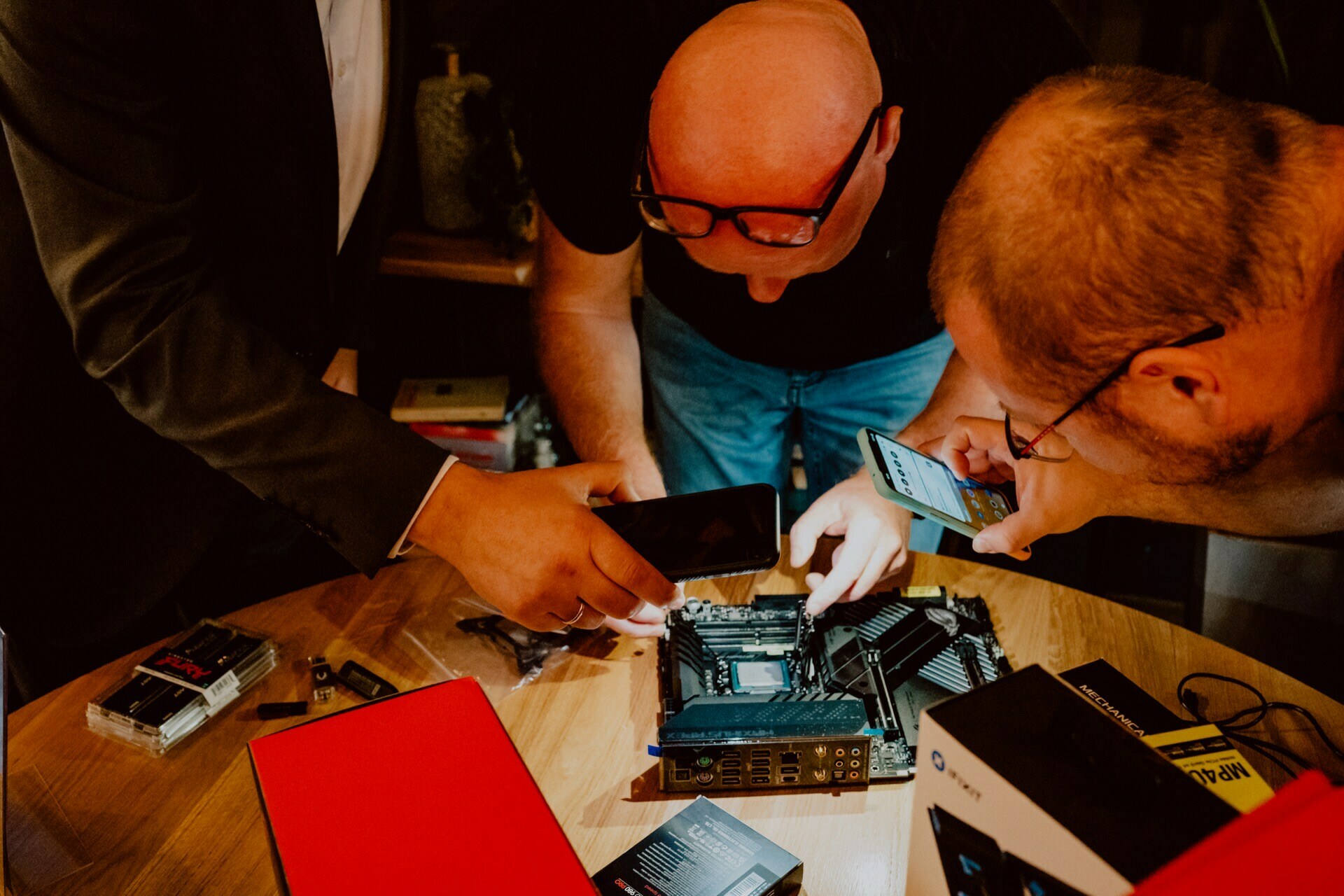 Three people are gathered around a wooden table, looking at a computer motherboard and other hardware components. Two of them are using smartphones, perhaps to document or examine the hardware, reminiscent of a press briefing scenario, while the third person points to the motherboard. 