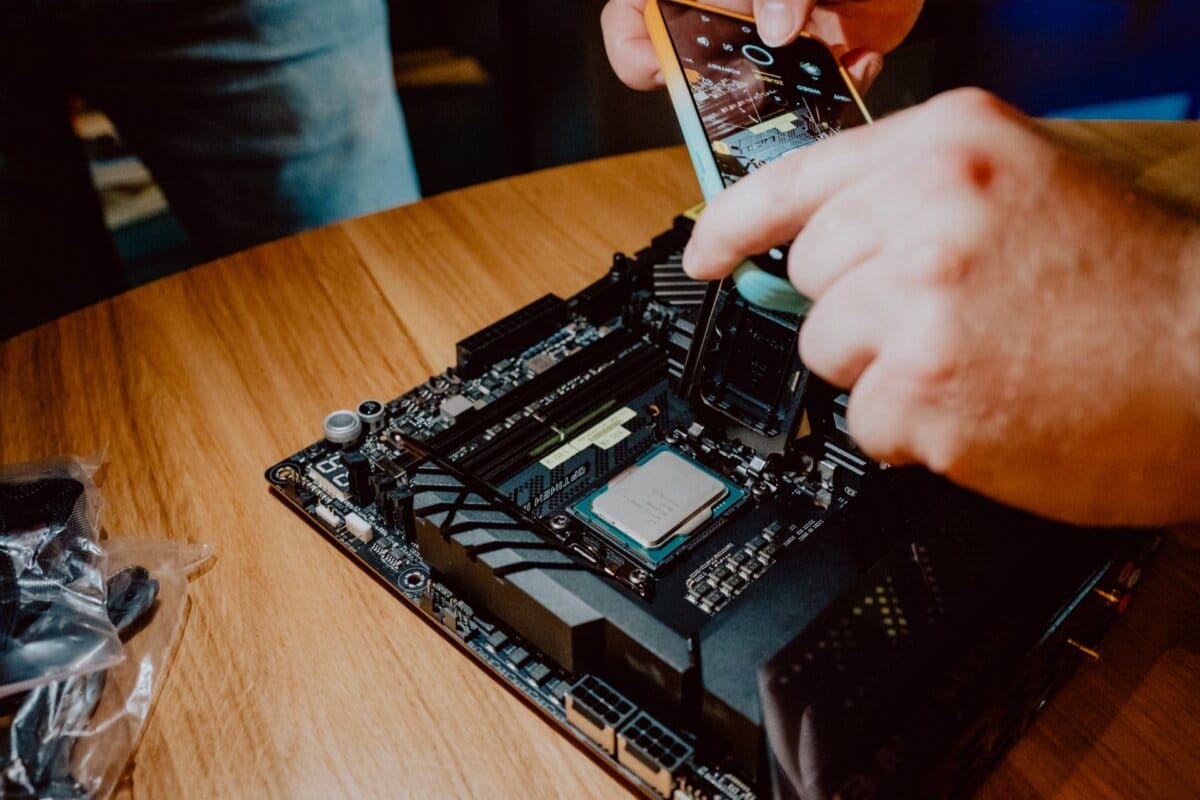 fotorelacja premiera procesorow Intel w LAS 65 Osoba używa smartfona, aby zrobić zbliżenie płyty głównej komputera na drewnianej powierzchni. Płyta główna jest częściowo zmontowana z widocznymi komponentami i obwodami. W pobliżu widoczne są ręce innej osoby, sugerujące współpracę lub pouczenie – być może w ramach spotkania prasowego.