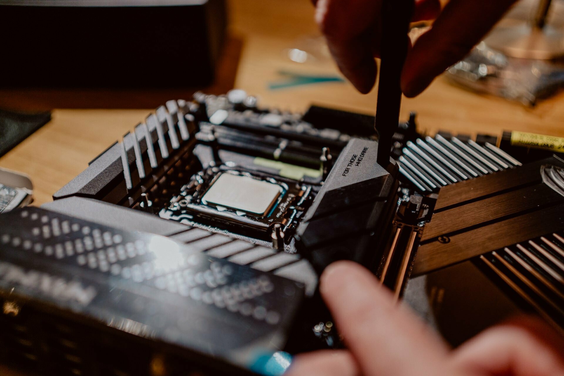 Close-up of hands assembling a computer motherboard. The photo focuses on the hands of a person using tools to attach components to a board placed on a wooden surface. Various electronic parts can be seen in the background, reminiscent of meticulous preparations for a press meeting.  