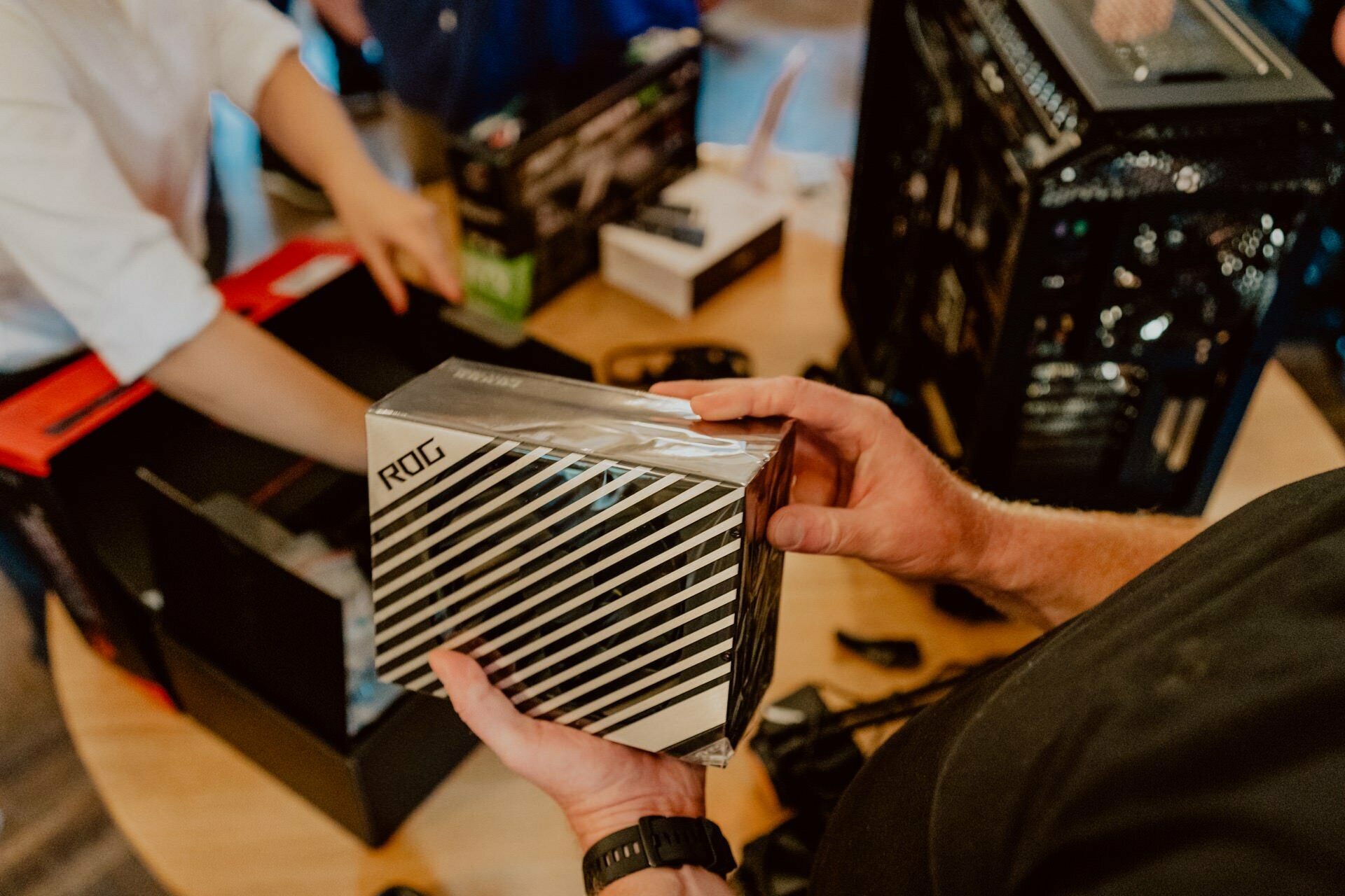 The person assembling the computer is holding a box with the ROG logo on it, as if preparing for a press meeting. On a table in the background, several other computer components and toolboxes can be seen. Another person is helping with assembly, and his hands are working on a component.  