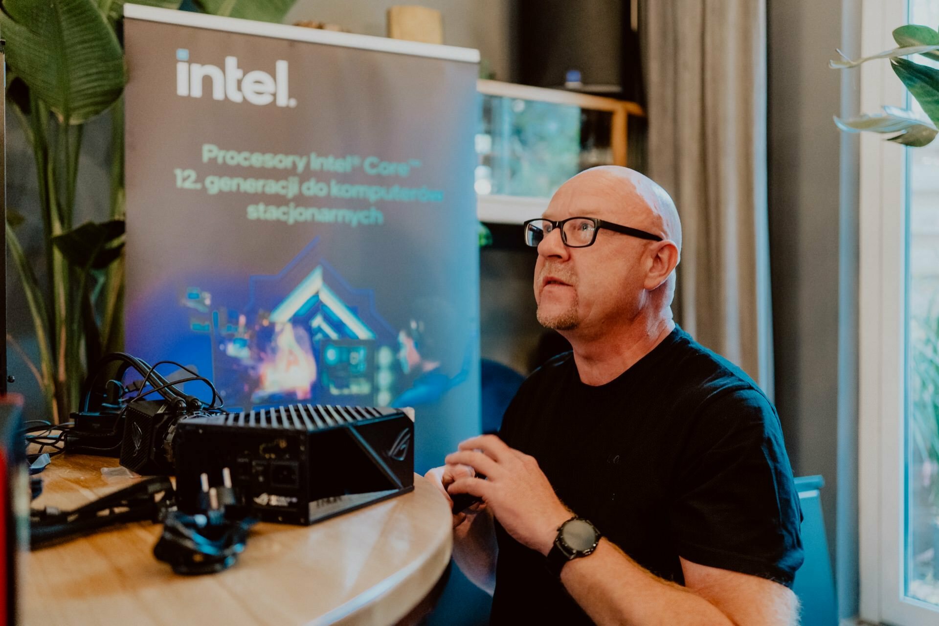 A person wearing glasses and a black shirt sits at a table with electronic equipment that looks like a computer component. Behind them, a banner with Intel's logo and Polish text about Intel Core desktop processors, referring to a press meeting. 