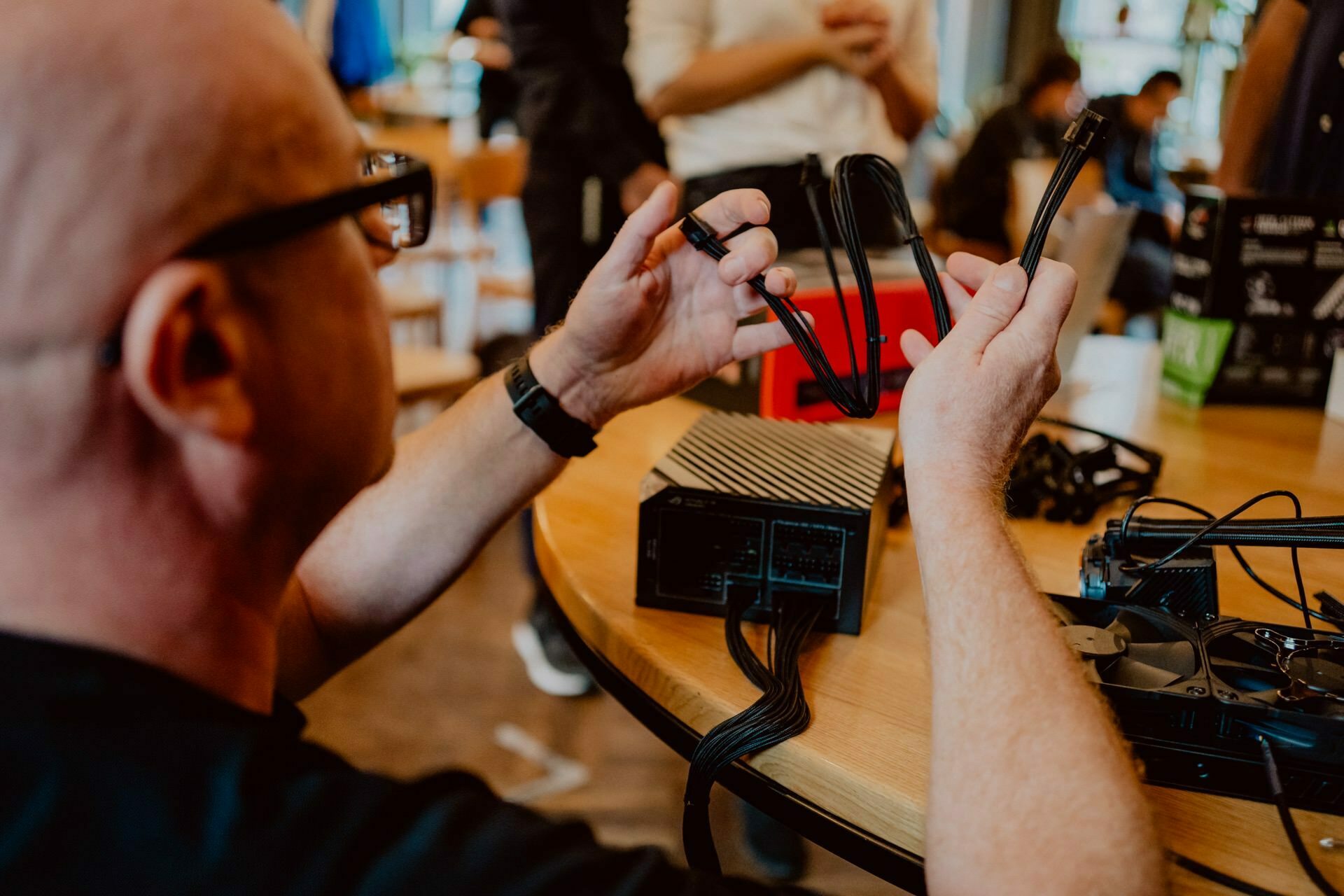 A man sits at a table and installs a power supply in a computer case. He is holding cables and connecting them to the device. The scenery resembles a technology workshop or press meeting, with other people and equipment visible in the background.  