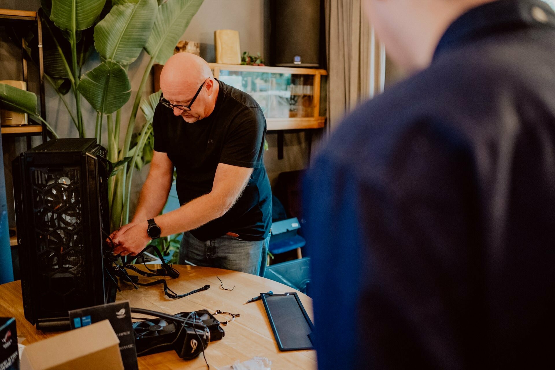 A man working on assembling a computer at a table on which various tools and components are spread out, as if preparing for a press meeting. He is concentrating on connecting wires inside the computer case. Another person is standing nearby watching him. A large plant and a cabinet can be seen in the background.   