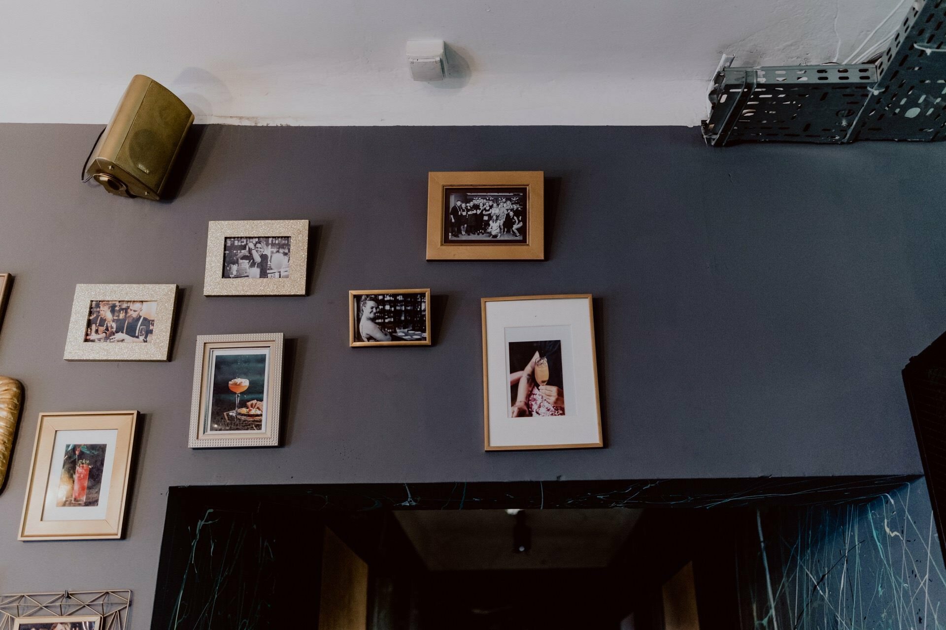 A wall with a collection of framed photographs and paintings of various sizes and orientations. The photos on a dark gray background depict scenes from photojournalism events. The speaker is mounted in the upper left corner of the wall.  