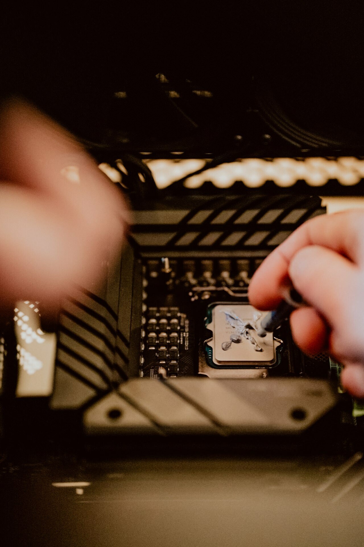 Close-up of hands applying thermal paste to a processor inside a computer, reminiscent of a press briefing. The processor is placed on a motherboard, surrounded by dark metal parts, highlighted by lighting. A person uses a syringe to spread the thermal paste evenly.  