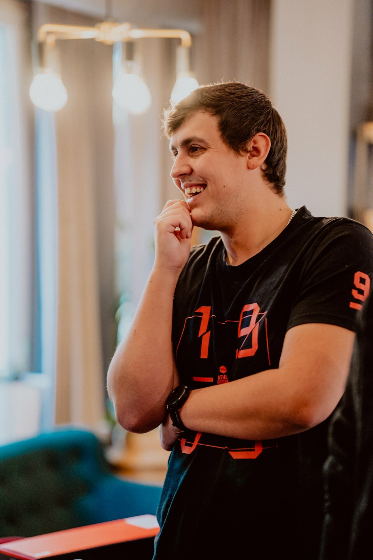 A person with short dark hair, wearing a black T-shirt with red numbers, smiles and stands with his hand resting on his chin in a brightly lit room during a press meeting. Blurred lights and curtains can be seen in the background. 