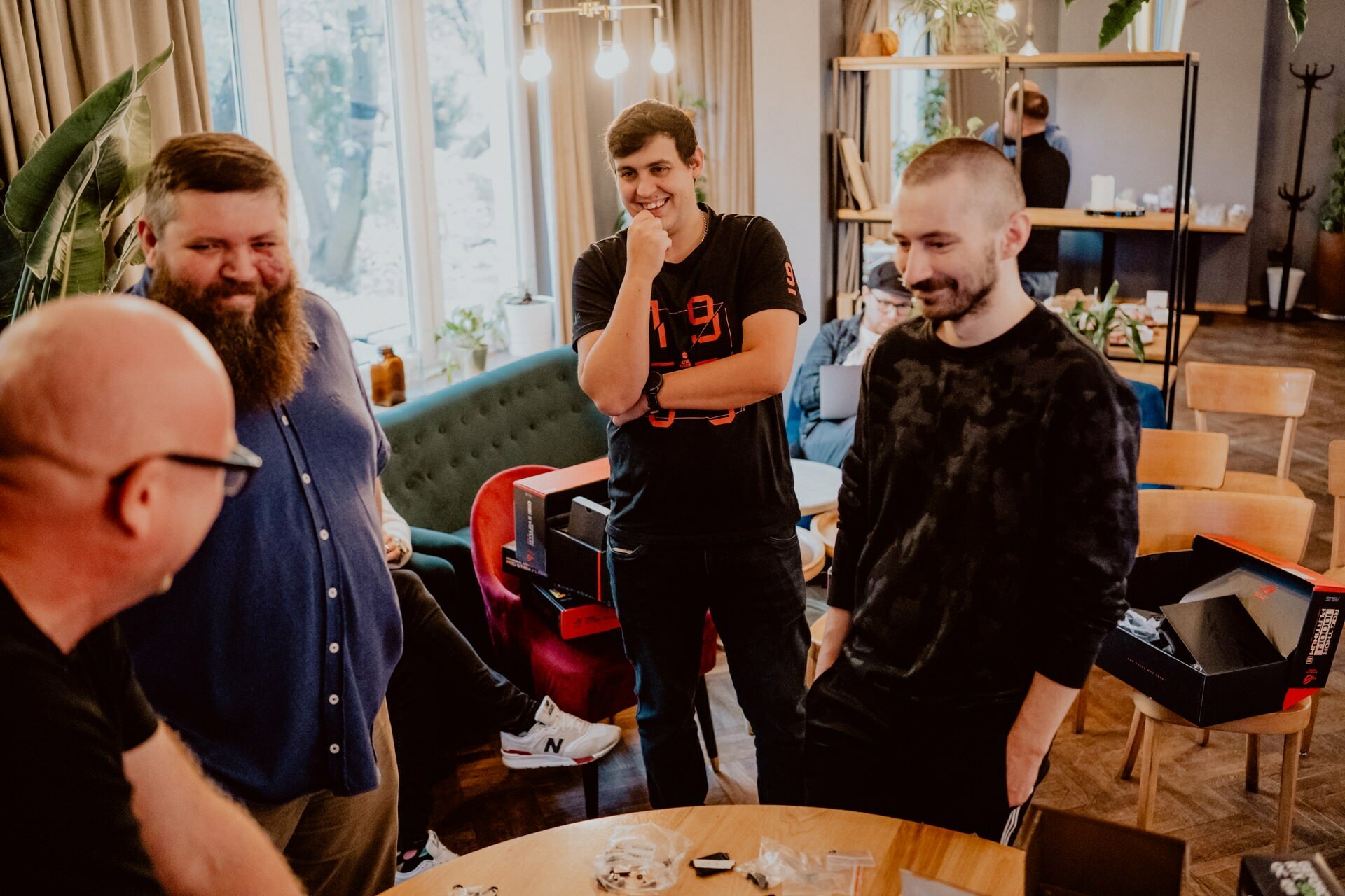 Four men are standing and smiling in a cozy room with wooden furniture and large windows, as if at an informal press meeting. One man is in a blue shirt, another in a black t-shirt and the third in a dark long-sleeved shirt. They appear to be having a carefree conversation at a table with some electronic gadgets.  