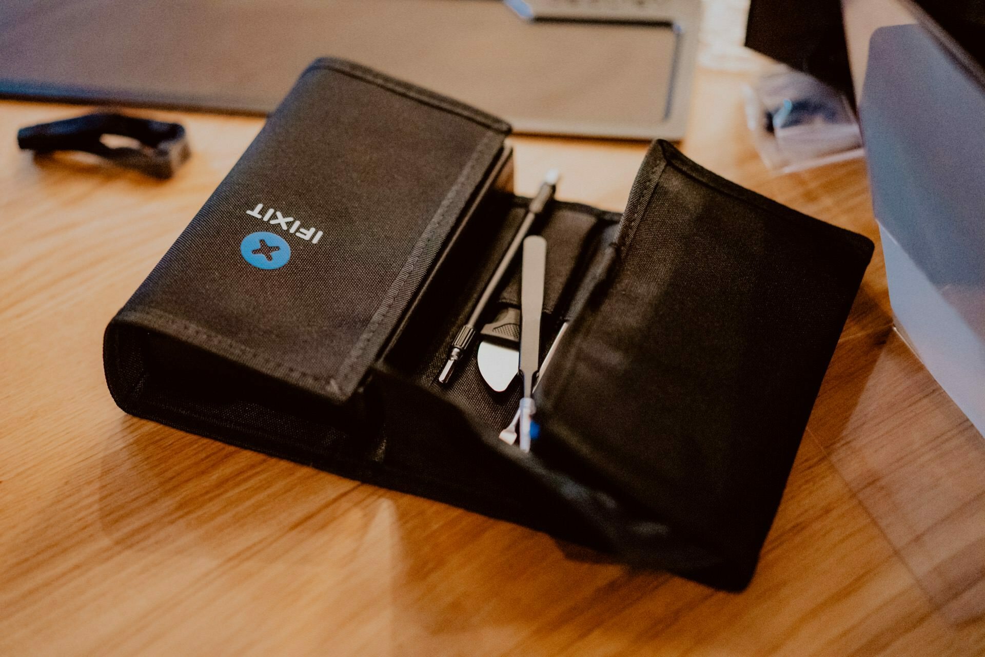 The black iFixit tool roll is open on a wooden surface and shows various precision tools neatly arranged in compartments, ready for the next press event. The outside of the roll shows the iFixit logo and a blue circular emblem with a white "X." 