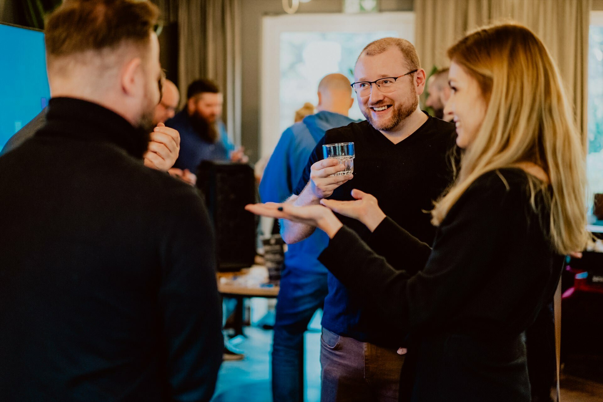 The three are having a conversation in a social atmosphere reminiscent of a lively press meeting. They appear to be smiling and enjoying their drinks. The background shows more people interacting, suggesting a lively atmosphere in a room with large windows letting in natural light.  