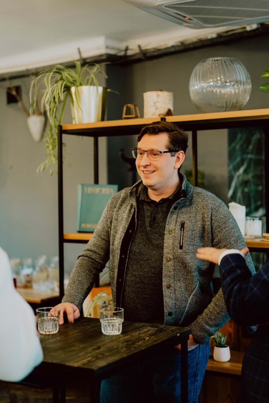 Osoba ubrana w szary sweter i okulary uśmiecha się i stoi przy małym drewnianym stoliku z dwiema szklankami wody, pozornie na spotkaniu prasowym. W tle znajdują się półki z roślinami i elementami dekoracyjnymi, a dwie częściowo widoczne osoby prowadzą z nimi rozmowę.