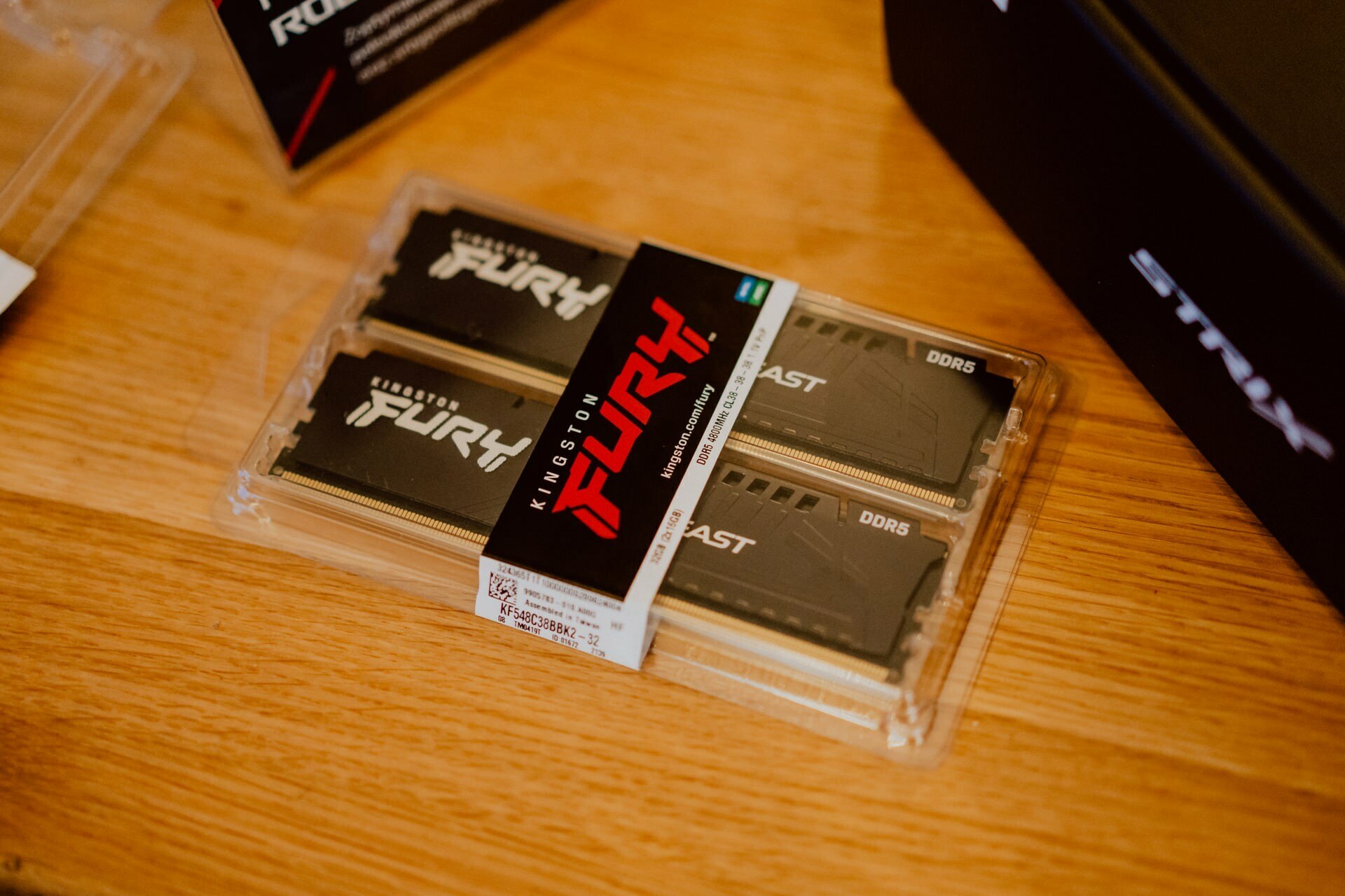 The package of Kingston FURY Beast DDR5 memory modules is placed on a wooden surface. On the package are two RAM dice with the "Kingston FURY" logo. Nearby, as after the press briefing, in the background you can partially see boxes with other computer components.  