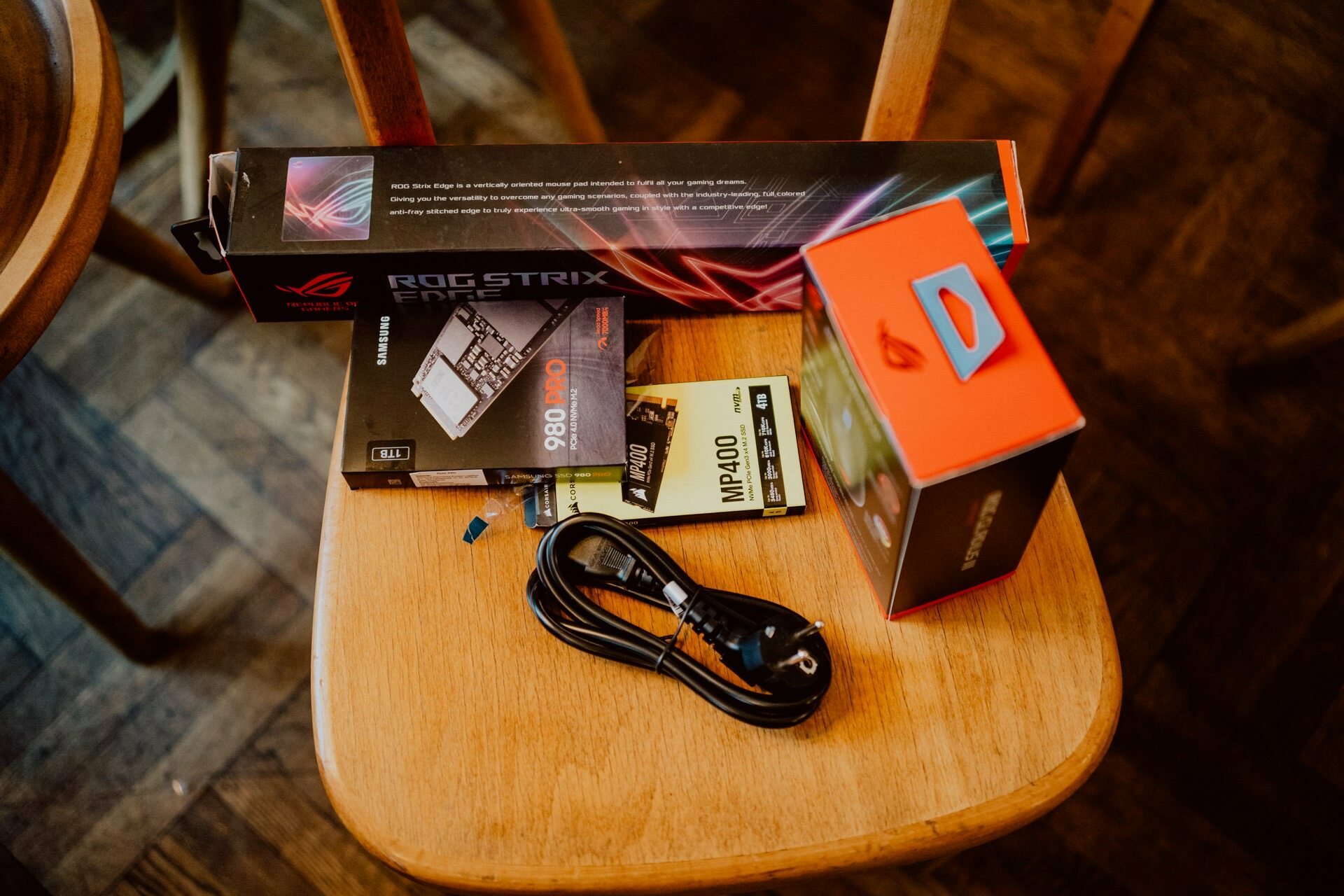 A collection of hardware components on a wooden chair, including an ASUS ROG Strix motherboard box, an MSI MPG series power supply box, a Kingston NV1 500 GB SSD box, a Corsair MP400 SSD box, and a black power cable - reminiscent of a technology enthusiast press meeting.