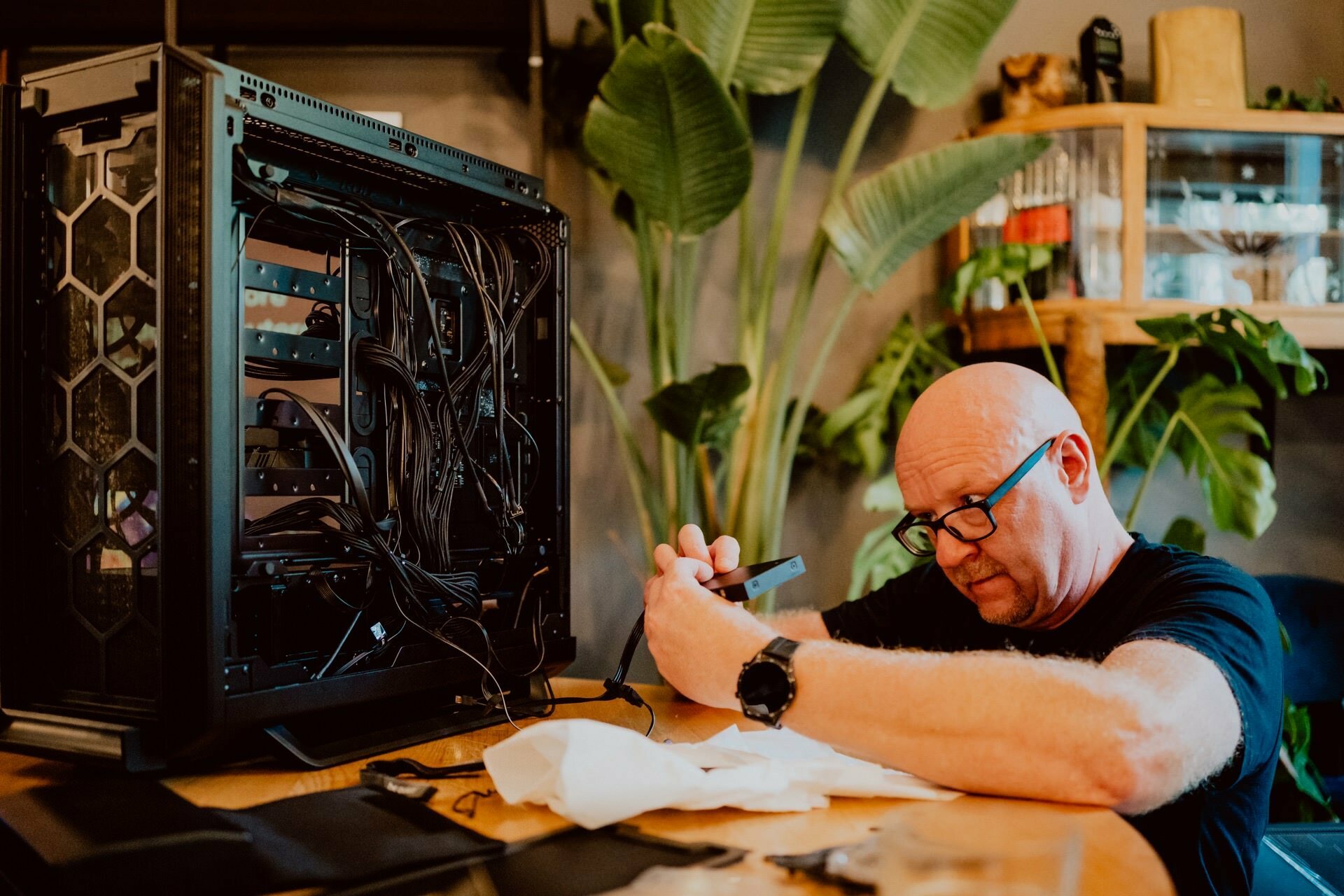 A bald man with glasses is busily working on assembling or repairing a computer. He is surrounded by tools and parts, and the open computer case shows numerous wires. In the background are leafy green plants and wooden shelves, as if preparing for a press meeting.  