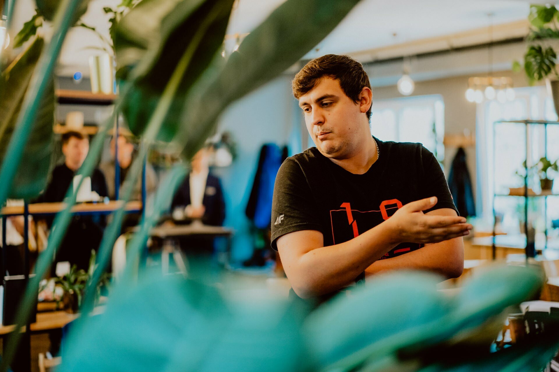 A man wearing a black T-shirt gestures with his right hand. He is standing in a room surrounded by large green plants. A blurry background shows other people talking, suggesting the casual and relaxed atmosphere of a press meeting.  