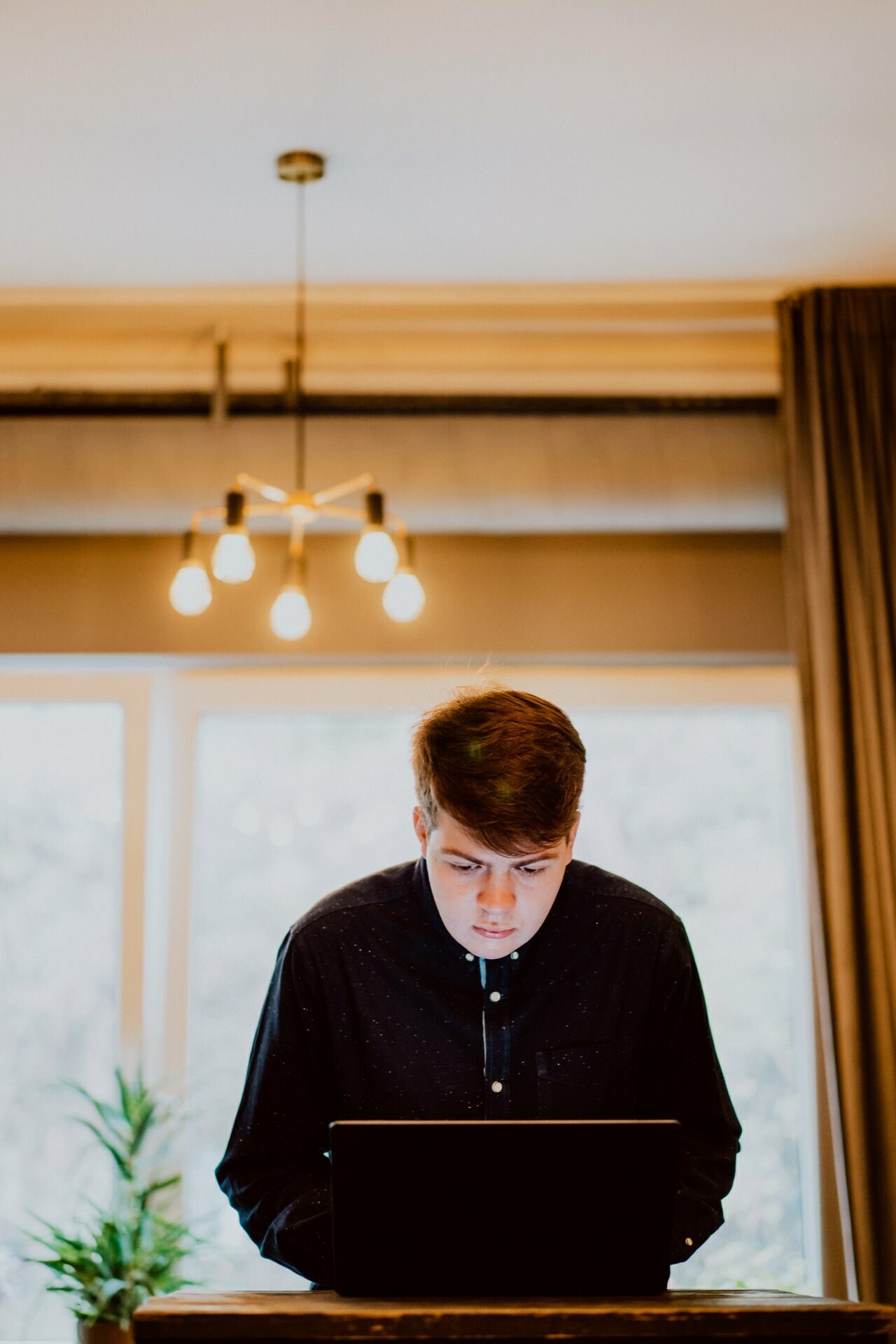 A young person in a dark shirt stands focused on a laptop at a desk in a well-lit room with large windows and curtains, preparing for a press meeting. Above them a modern pendant light fixture with five bulbs is visible, casting a perfect glow. On one side of the desk stands a small plant.  