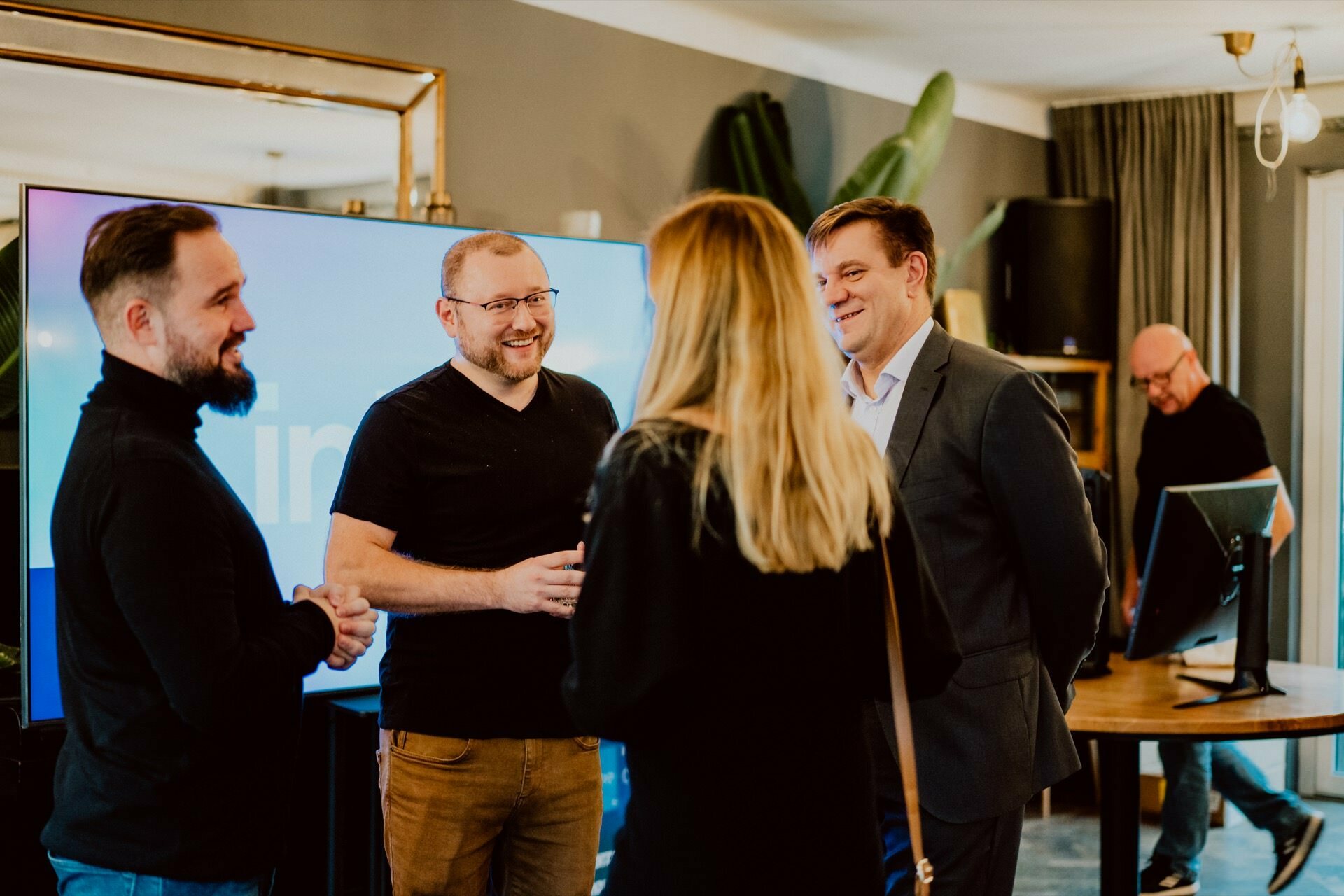 A group of five people are engaged in a lively conversation in a room. Four are standing in front of a large screen and a reflective frame, reminiscent of a press meeting; one person is working at a table in the background. The environment seems casual but businesslike.  