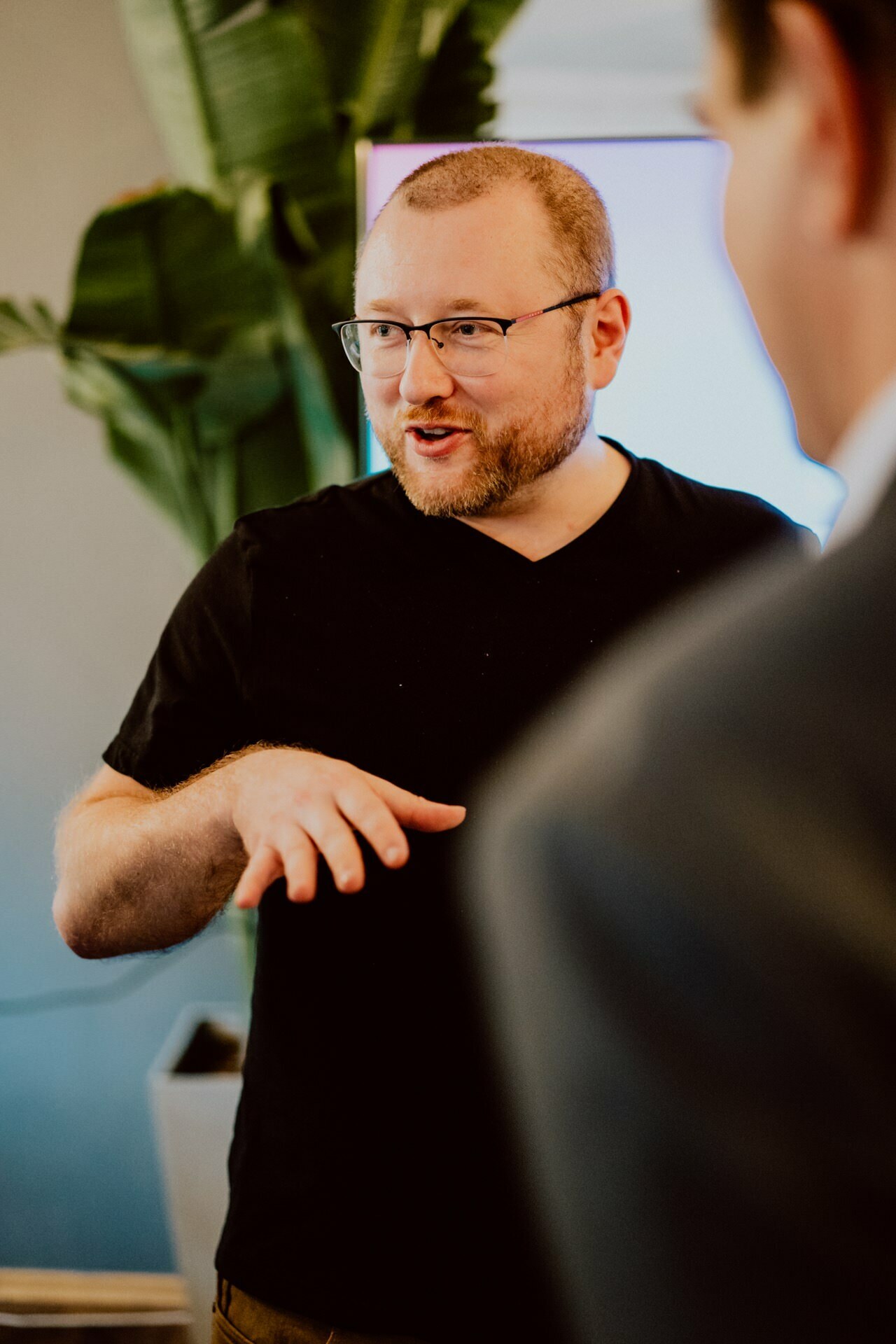 A man wearing glasses and a beard is having a casual press conversation. He is wearing a black T-shirt and gesturing with his hands. The background includes a large green plant and a vague person facing him, creating a casual atmosphere.  