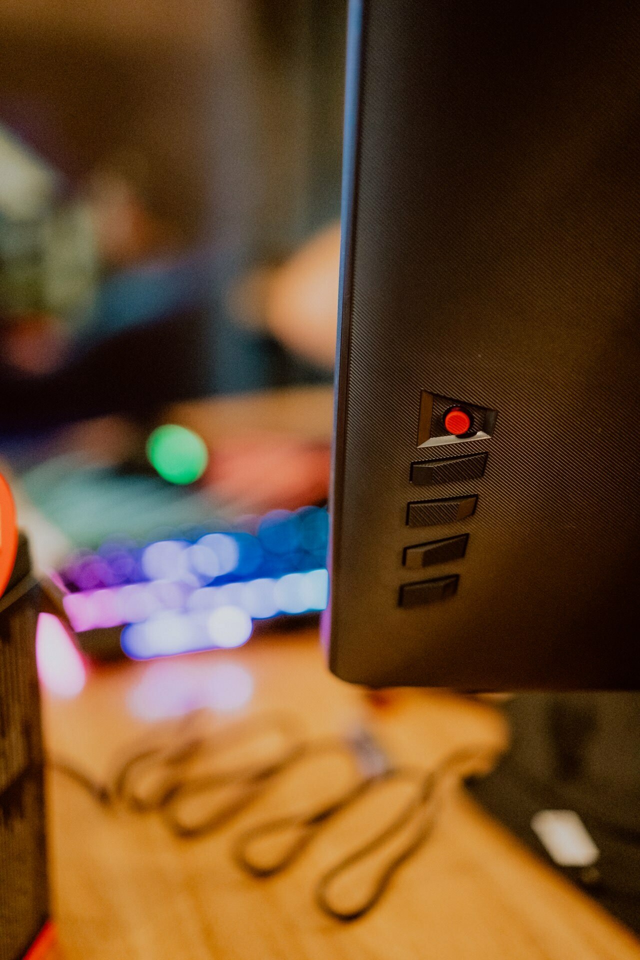 Close-up of the back of a computer monitor showing a red power button. The background is blurred, showing a colorful backlit keyboard on a wooden desk, reminiscent of the setup at a press briefing, and some out-of-focus elements on the left. 