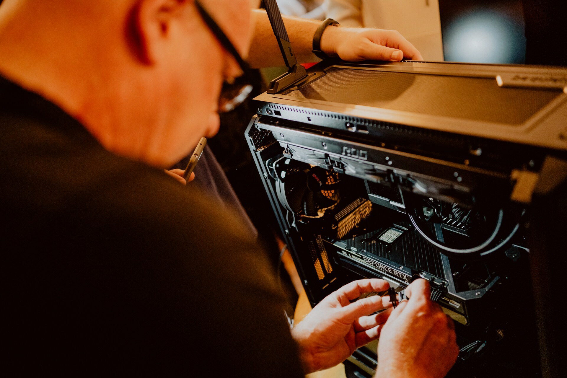 A person wearing glasses and dark clothing works inside the computer tower, focusing on its components. Another person's hand holding a tool helps from above. The scene is lit with warm light, reminiscent of an intimate press meeting.  