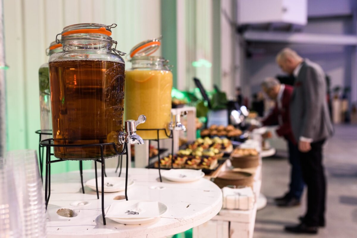 Glass jars full of juices and in the background men selecting snacks from the catering table