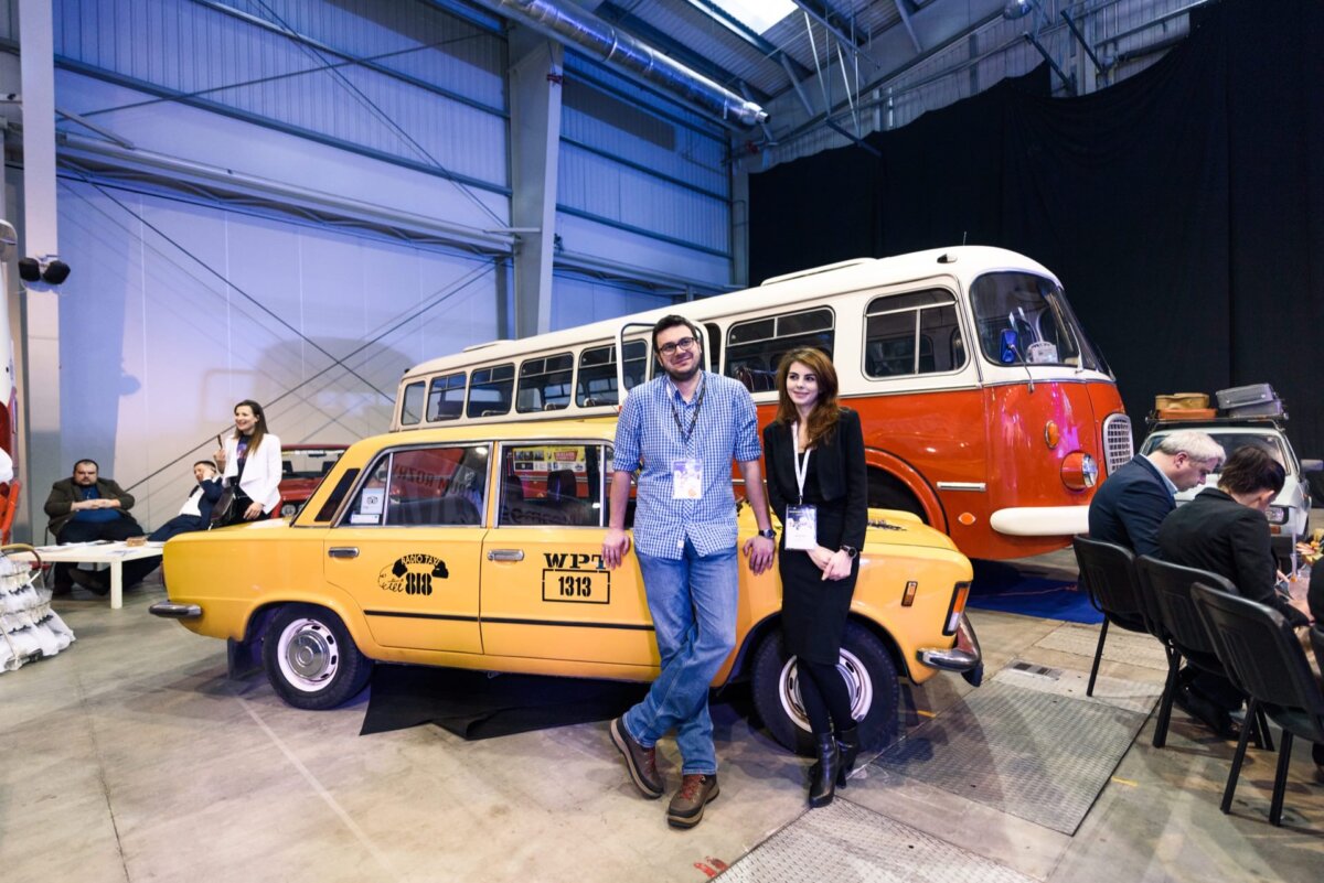 Girl and boy leaning against an old yellow WTP cab and in the background is a red bus called a cucumber