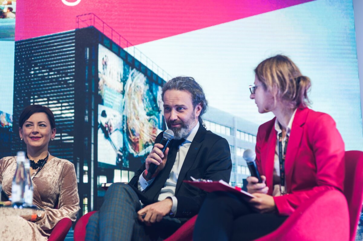 Man with beard speaks into microphone between two women