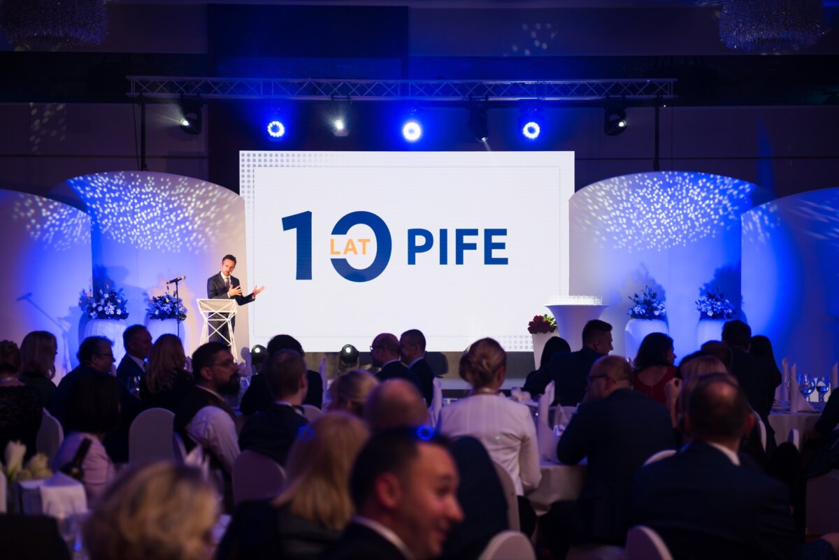 A person stands at a podium on a stage in front of a large screen that displays the words "10 YEARS OF PIFE." The event takes place in a dimly lit room filled with round tables where attendees are seated. Blue lighting and flower arrangements decorate the space, conveying an elegance perfect for event photography.  