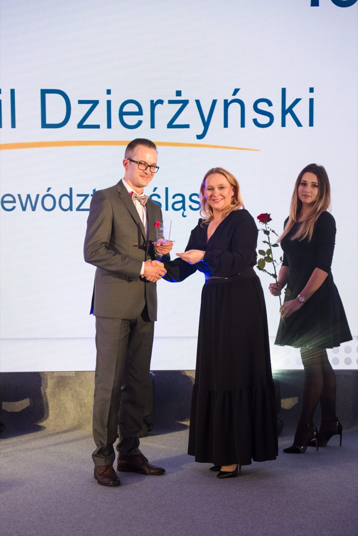 During the award ceremony, a man in a suit holds an award, shaking hands with a woman in a black dress, who also holds a certificate. To the side stands another woman, also in a black dress, holding a red rose. This moment was captured perfectly by the event photographer warsaw.  