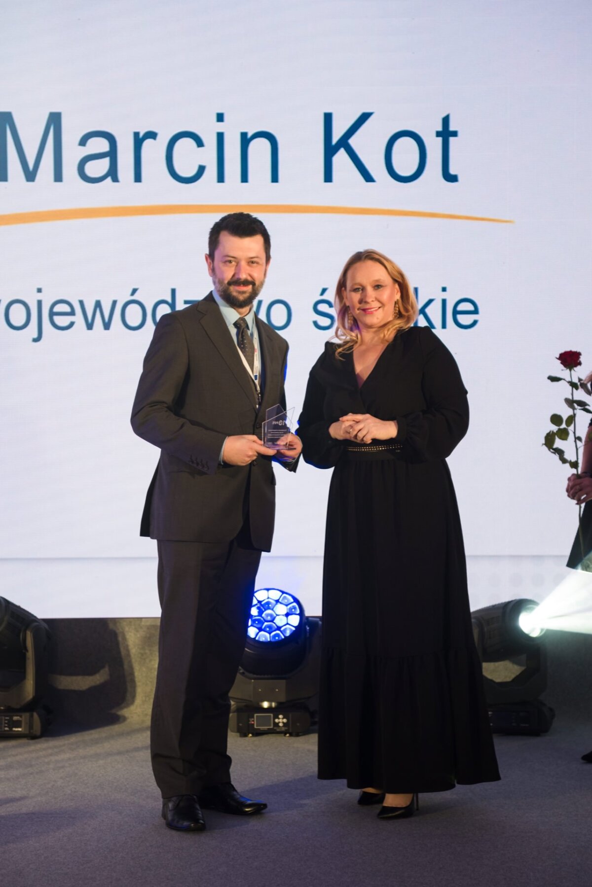 A man and a woman stand on stage, holding an award. The man is dressed in a suit and tie, the woman in a long black dress. Both are smiling at the camera, behind them is a large screen with the name "Marcin Kot" on it. Stage lights and a single rose are visible in this event photo.   