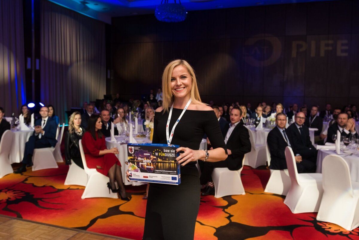 A smiling woman in a black dress holds a LEGO Friends set while standing in front of a seated audience at a formal event. Elegant decor with white chairs and a colorful floral rug adds to the charm. The large logo is partially visible on the wall behind her, capturing the essence of this event photography.  