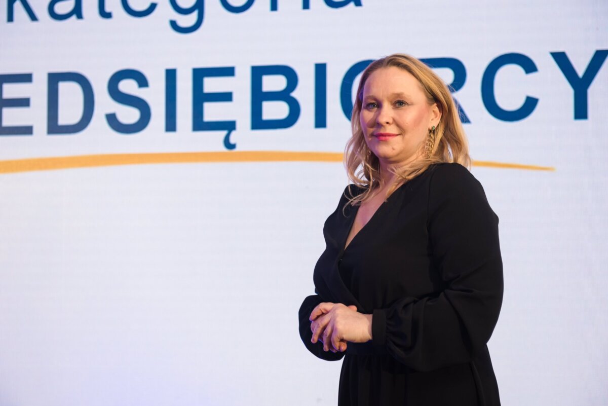 A woman with blonde hair is standing in front of a background with Polish text that reads "ENTERPRISE." She is wearing a black dress and an expression of confidence, which is perfectly captured in this event photo. 