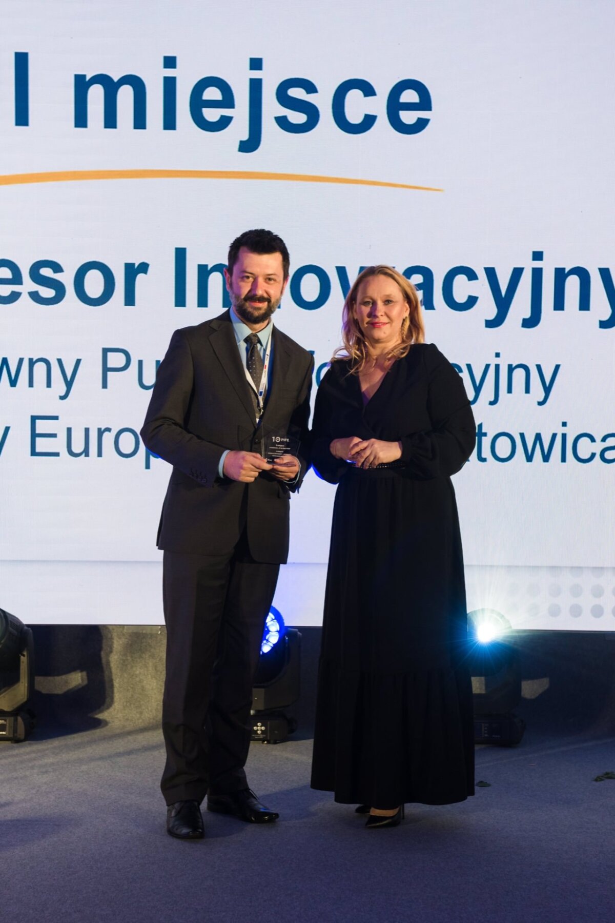 A man in a suit and tie and a woman in a black dress pose for a photo on stage, while behind them is a large screen displaying text in Polish. The man is holding an award plaque and both are smiling at the camera. Lighting and stage equipment can be seen in the background, captured by event photographer warszawa.  