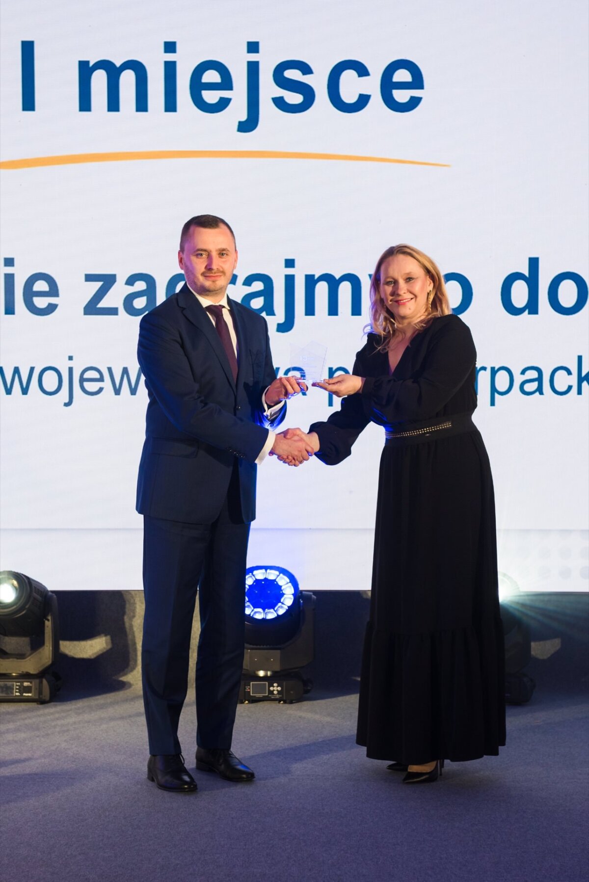 A man in a suit and a woman in a black dress are shaking hands on a stage with Polish text in the background, and stage lights illuminate the floor behind them. The two are smiling, capturing a moment that could be perfect for event photography or photo coverage of events at an awards or recognition gala. 