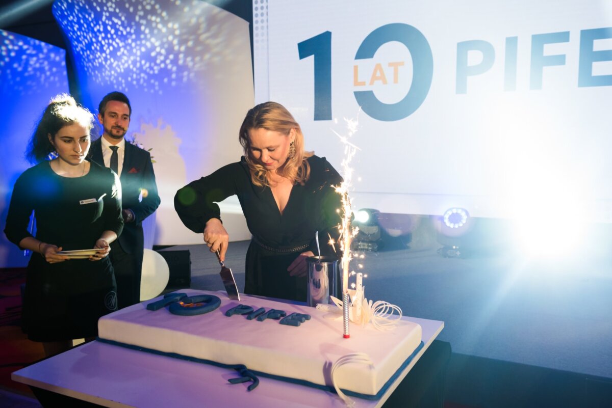 A woman cuts a large rectangular cake with a knife while a firework lights up on the cake. Two people stand nearby and watch. In the background is a large sign with the words "10 YEARS OF PIKE" illuminated by a bright light, which perfectly captures the moment of the photo-recording of the events.  