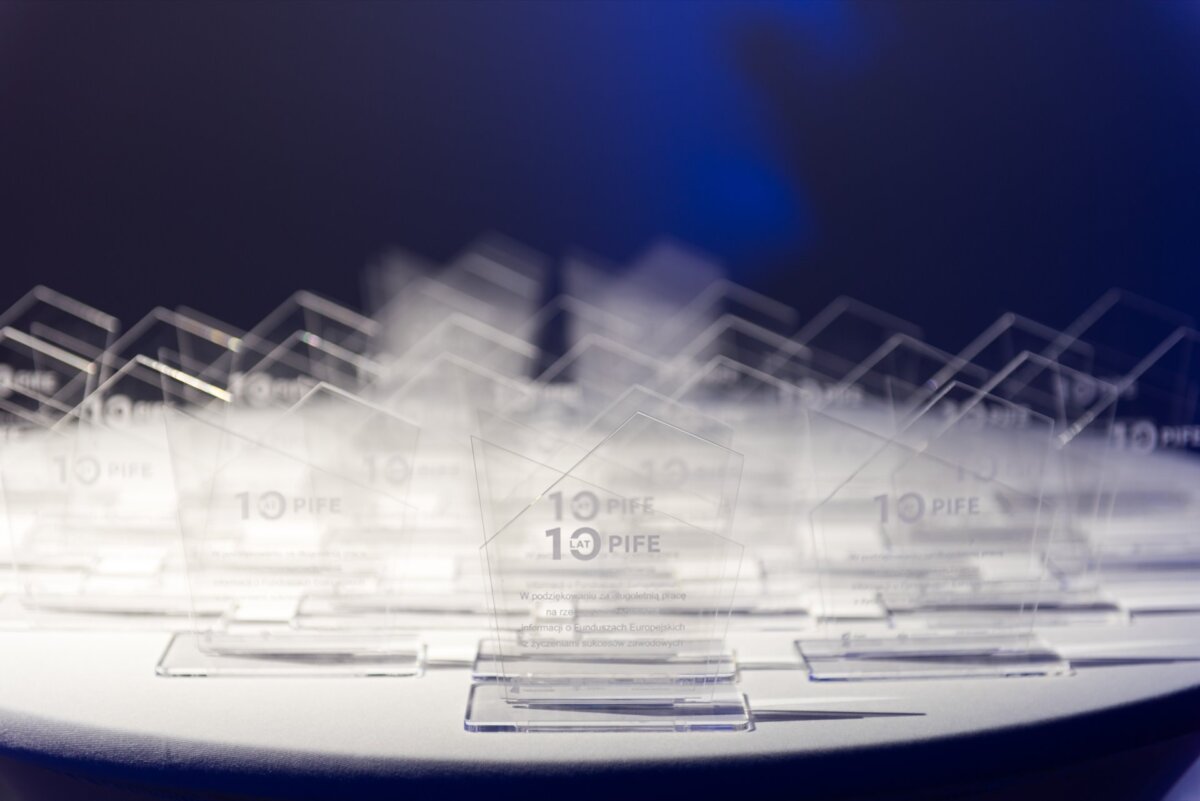 A collection of clear glass awards stands on a round table with a dark background. The awards are engraved and arranged in several neat rows, reflecting the light. The nearest award bears the inscription "10 PIFE", with additional smaller text engraved below, capturing the essence of event photography.  