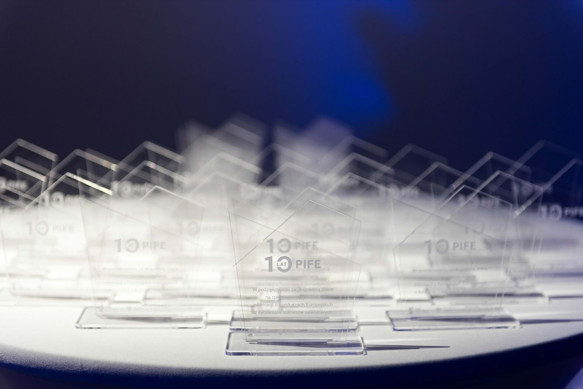 Reihen glänzender Pokale mit graviertem Logo, aufgestellt auf einem runden Tisch vor der Eröffnung der Gala, dunkler Hintergrund und blaues Licht.