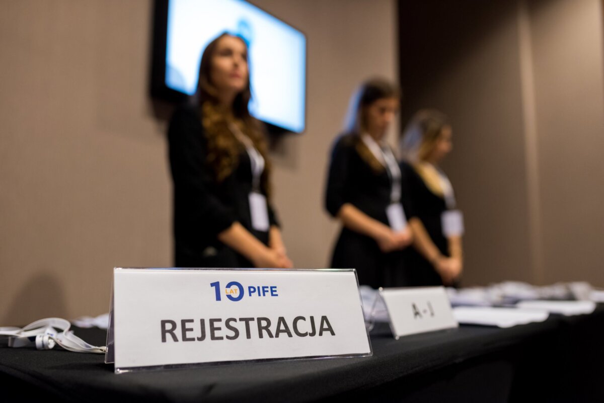 The license plate reads "REGISTRATION" with the PIFE logo, and in the background stand three women dressed in black and with badges. The room has a professional vibe, with a screen on the wall, perfect for capturing photo coverage of events by any event photographer warsaw. 