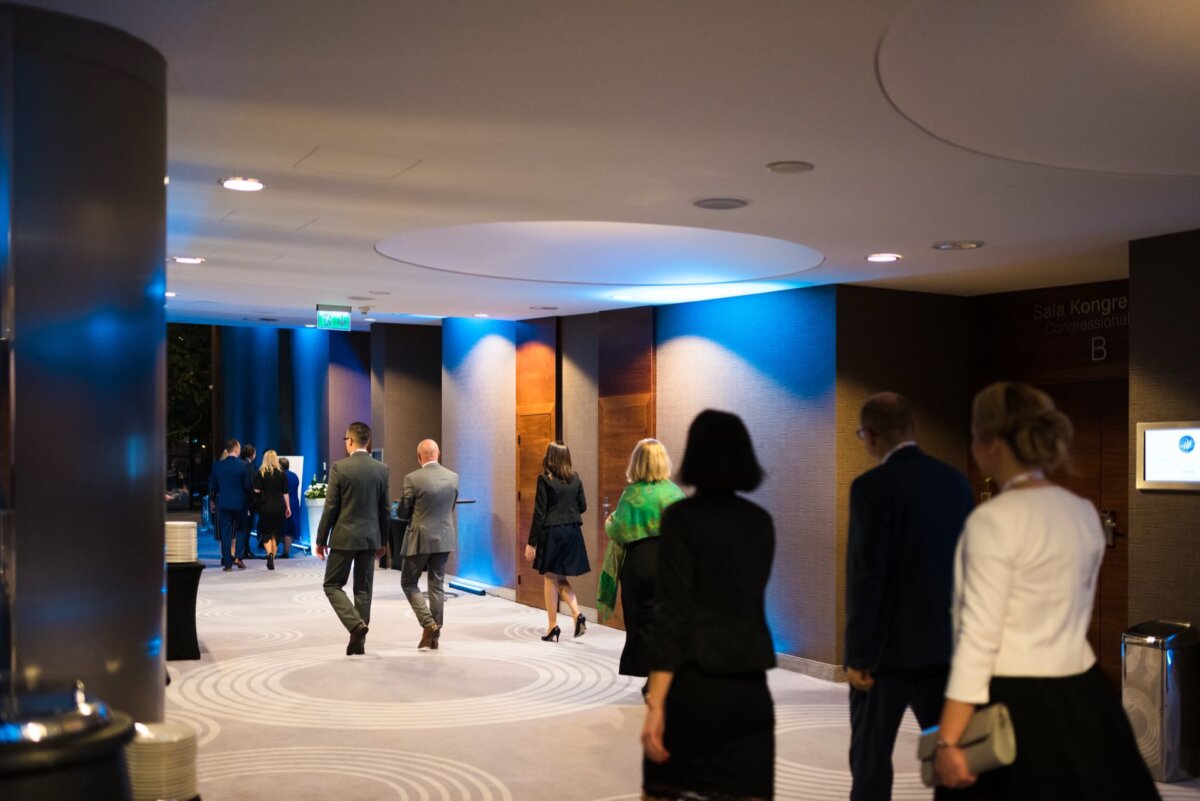 People dressed in business attire walk down a lighted corridor toward a conference or event room. The corridor is equipped with modern lighting, paneled walls and round ceiling fixtures. The atmosphere gives the impression of being professional and organized, perfectly suited for event photography.  