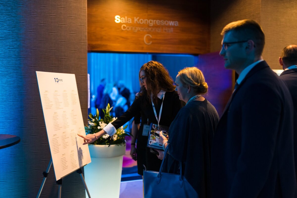 During the event, three people stand near a large blackboard. One woman, pointing to the board, makes a connection with the others, who are watching intently. Above, the board reads "Congress Hall" and "Congress Hall" with the letter "C." The background has a blue atmosphere reminiscent of professional event photography in Warsaw.   