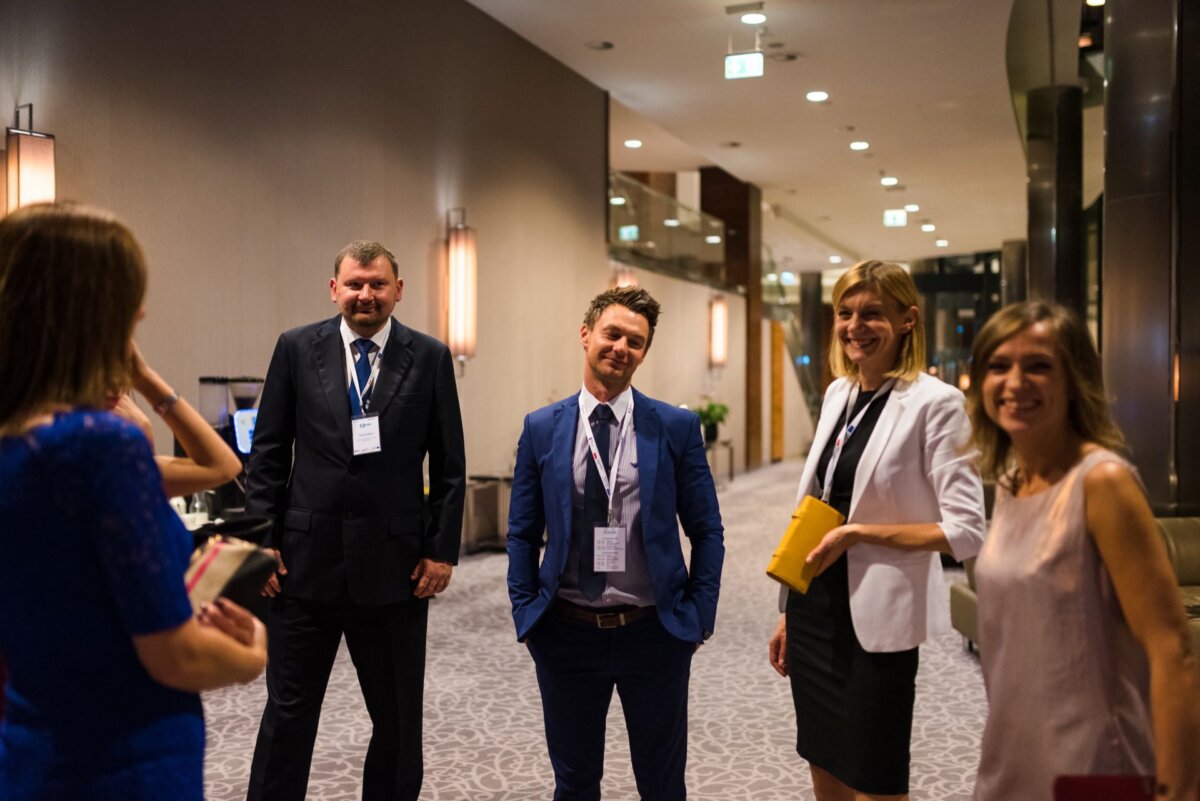 A group of five professionally dressed people stand in a well-lit, newly decorated hallway, immersed in conversation. The three men and two women, smiling and wearing event badges around their necks, appear to be attending an official gathering. This scene captures the essence of event photography.  