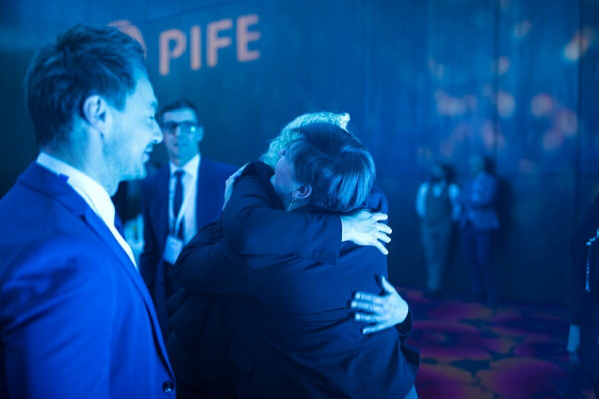 A group of people in business attire at an event taking place in a room lit by blue light. The focus is on two hugging people in the foreground, while other people look on and smile. The wall in the background partially shows the word "PIFE," beautifully captured by event photographer Warsaw.  